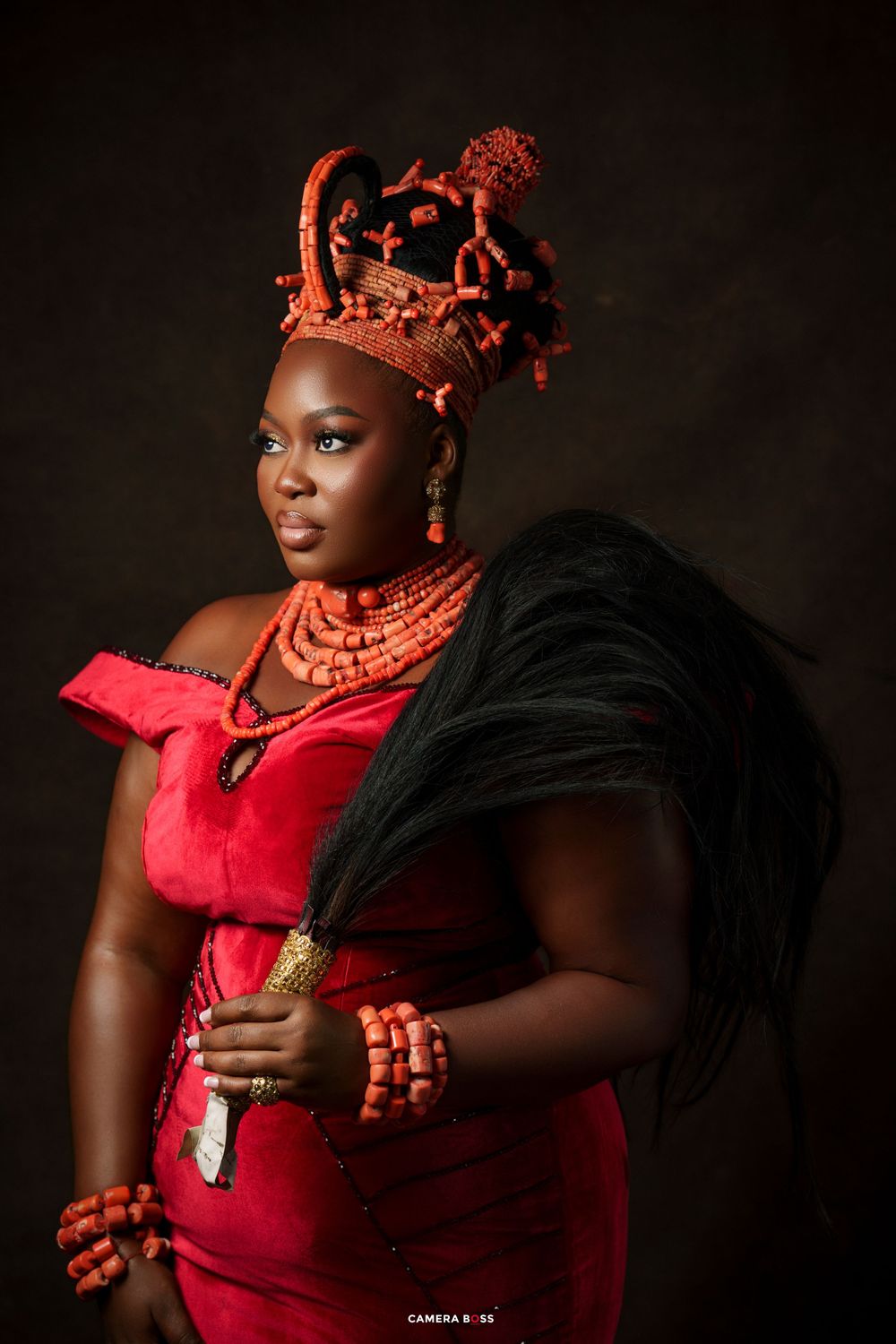 Woman in red velvet dress wearing traditional Nigerian coral beaded crown and jewelry in a regal portrait.