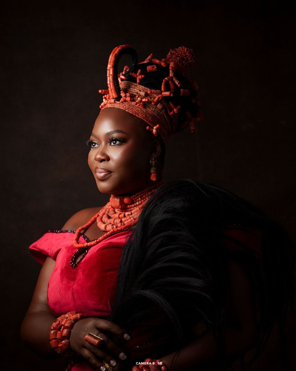 Woman in traditional Nigerian attire with red coral beaded crown, jewelry, and velvet dress against dark background.