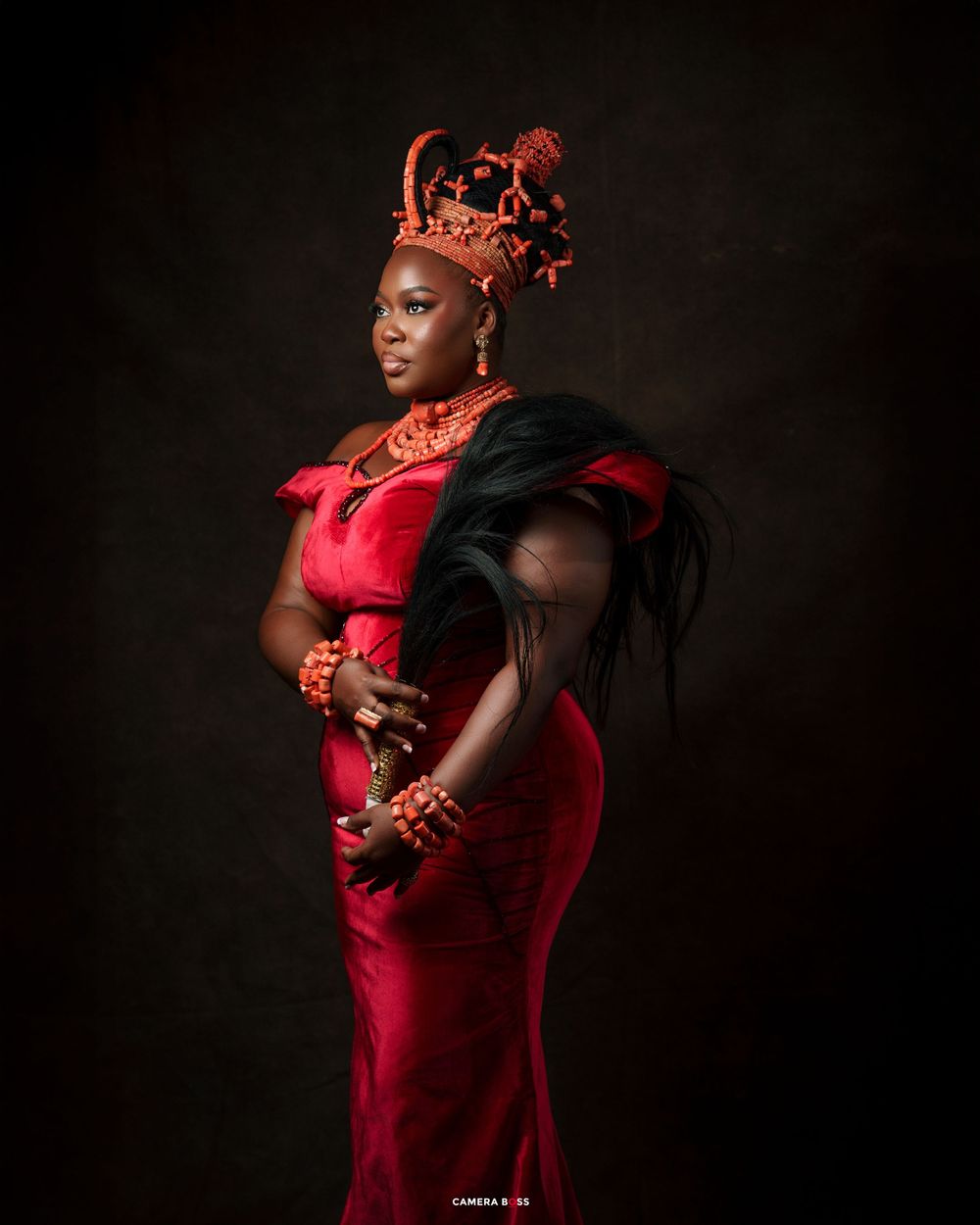 Woman in red velvet gown with coral beaded jewelry and ornate crown in traditional African royal attire.