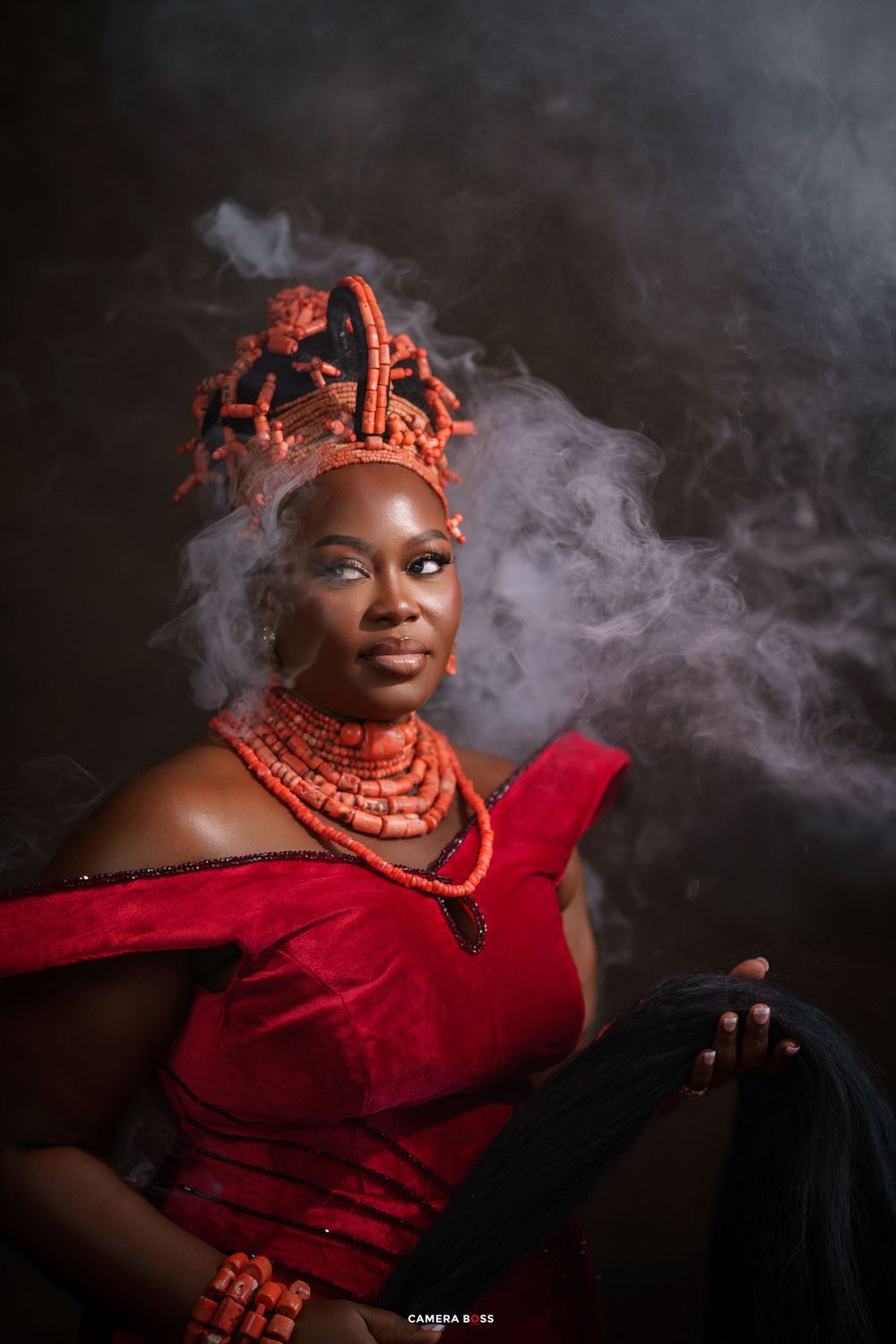 A regal African woman in red traditional attire with coral beads and ornate headdress poses dramatically with smoke swirling around her.
