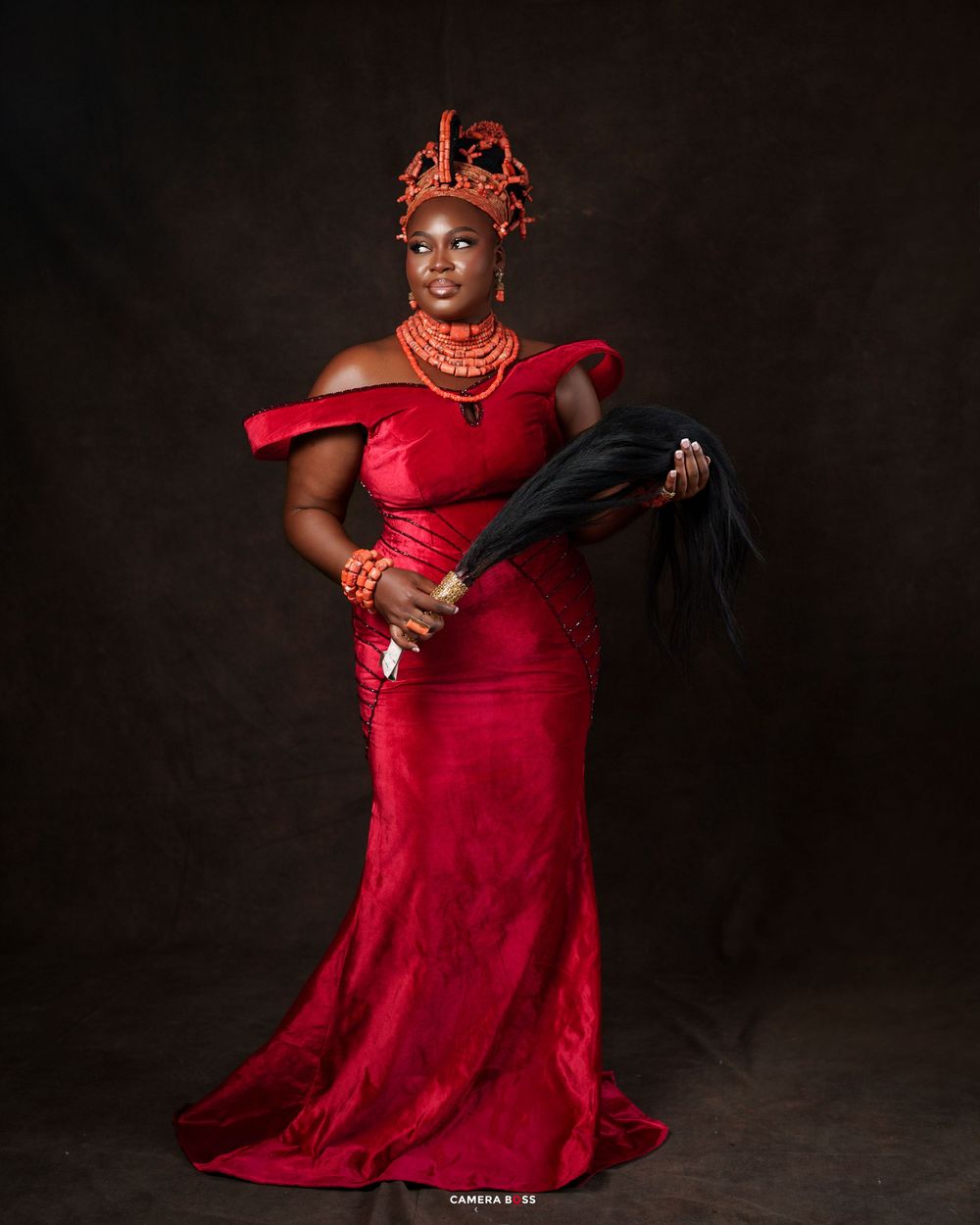 Woman in red off-shoulder gown with coral beads and headdress holding a black fan in a studio portrait.