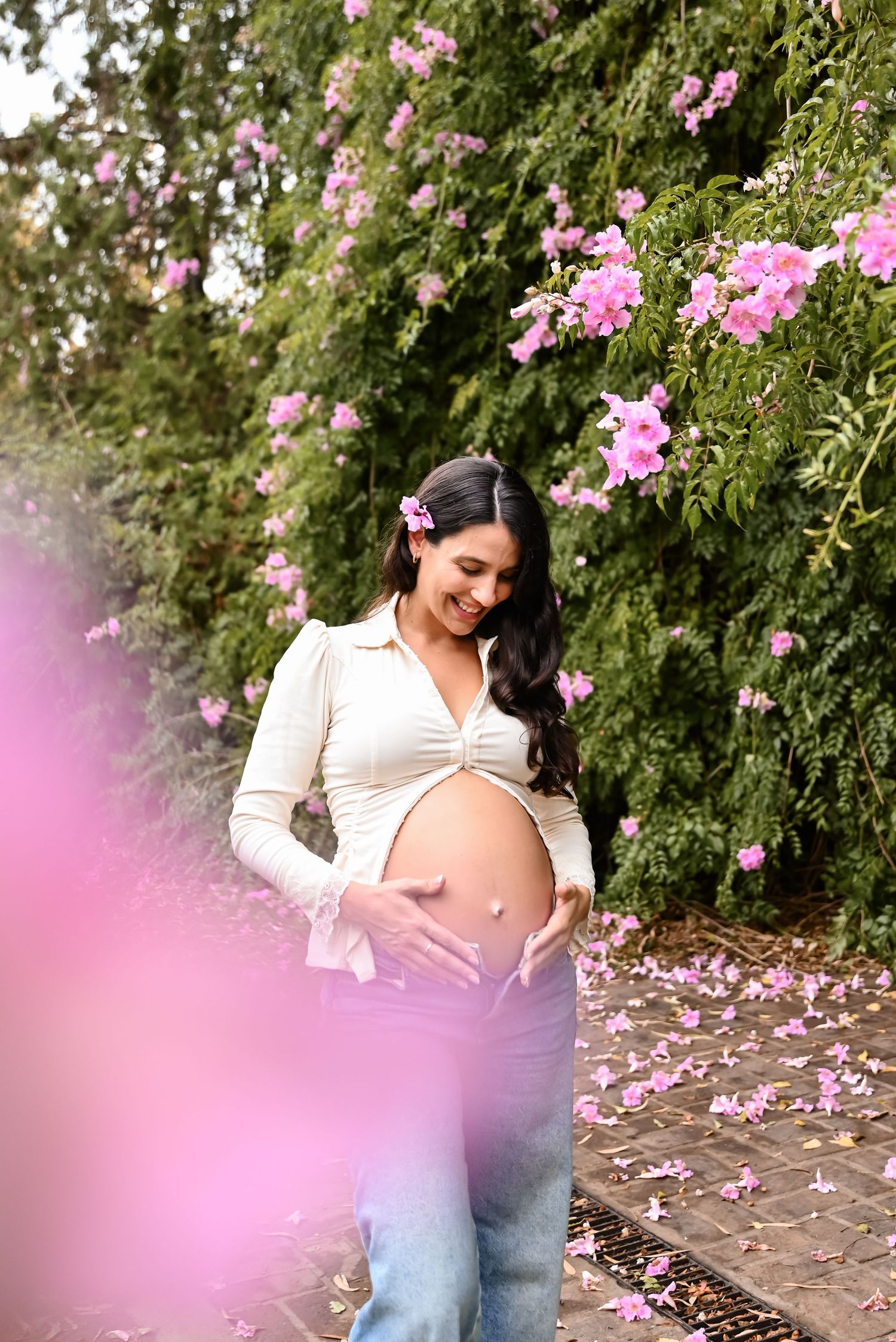 Smiling pregnant woman in white crop top and jeans cradling her baby bump outdoors near pink flowering vines.