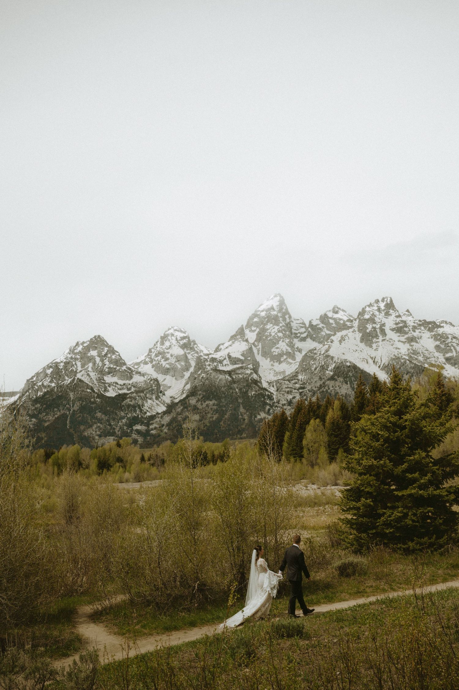 Bride and groom walk a mountain path with snow-capped Grand Teton peaks and lush green trees in the background.