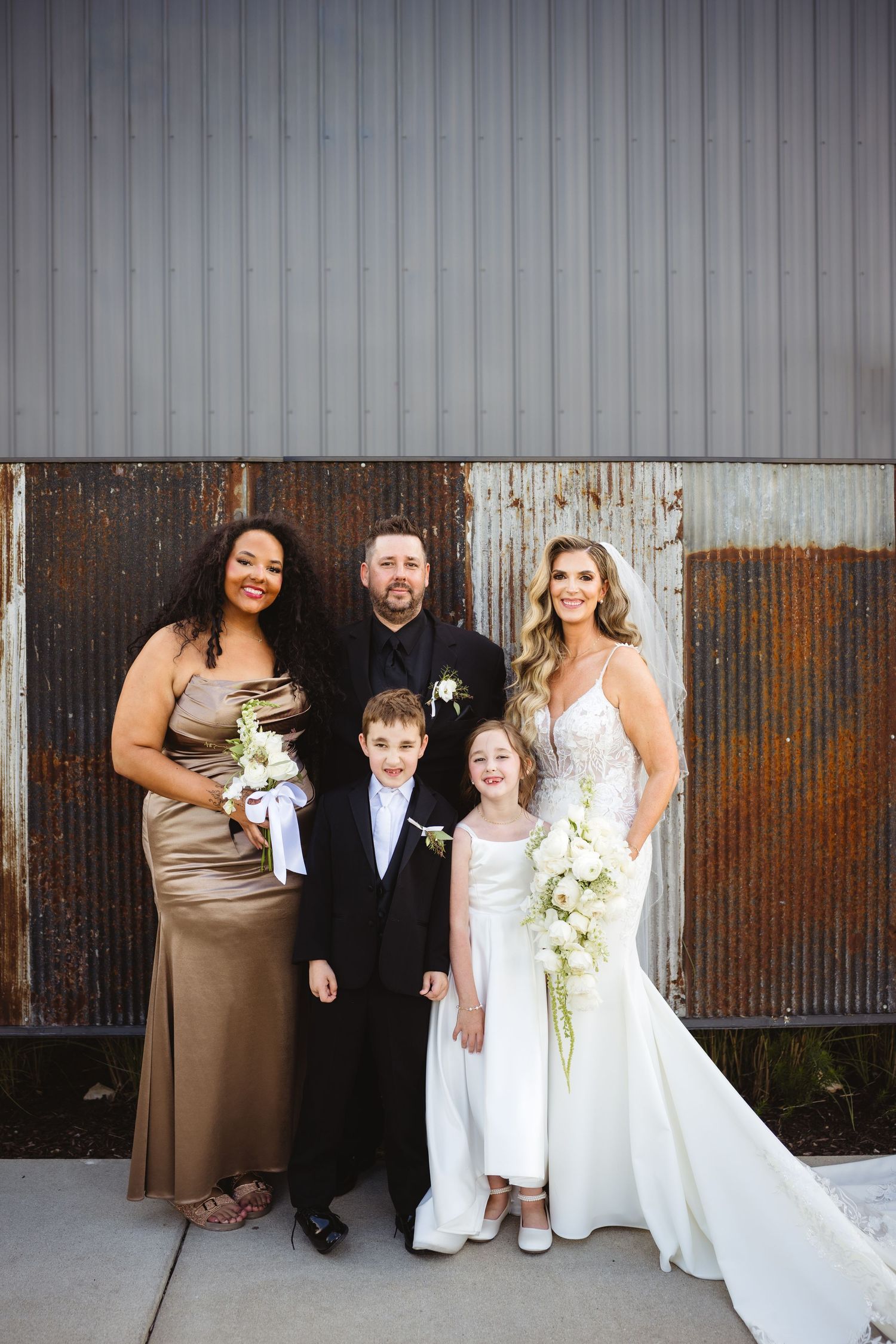 Wedding party poses before rustic metal barn wall, bride in white gown with bouquet, groom, bridesmaid, and two children.