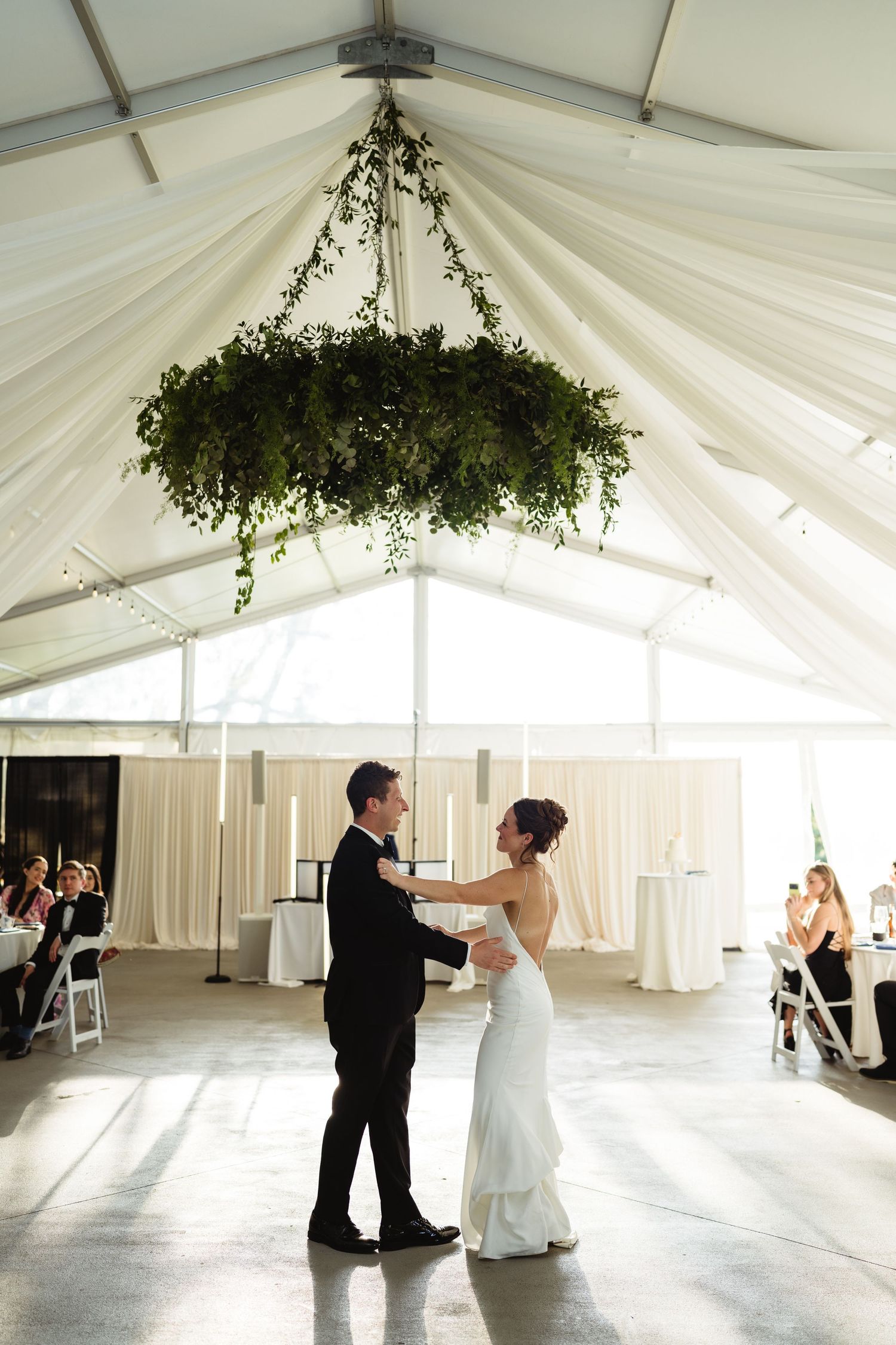 Bride and groom share first dance under greenery chandelier in elegant white draped wedding tent.
