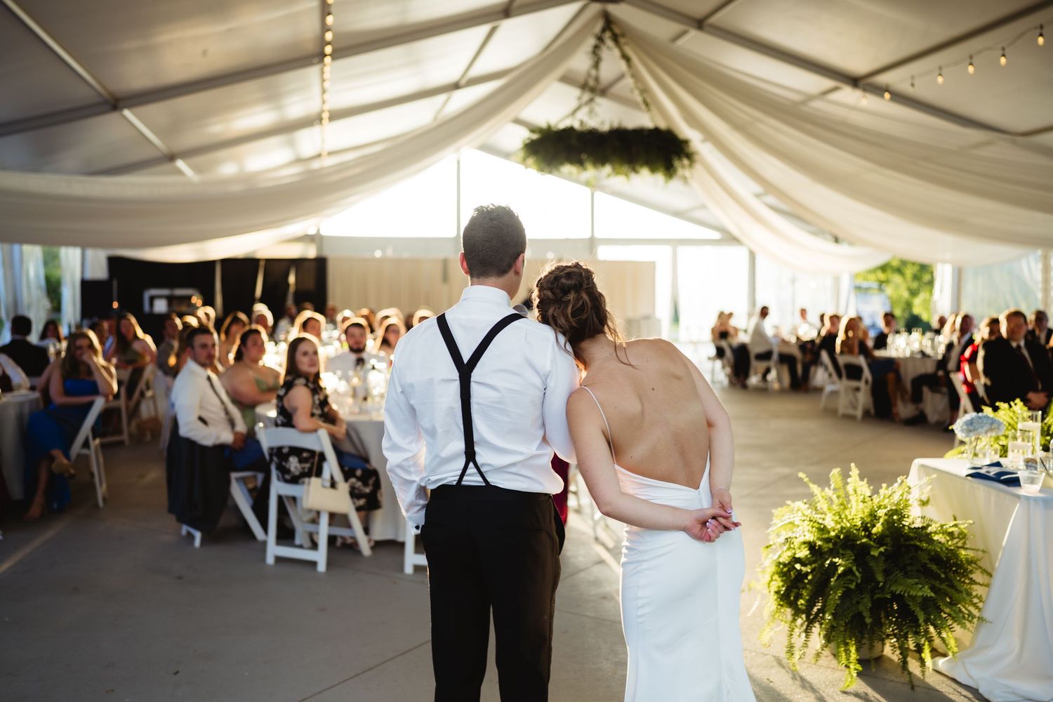 Bride and groom stand before guests at elegant tented wedding reception with draped ceiling and string lights.
