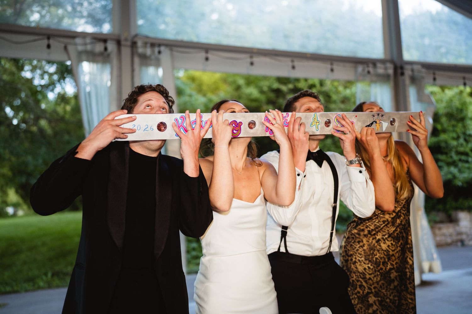 Wedding party holds pizza boxes in front of faces for fun photo at outdoor tented reception venue.