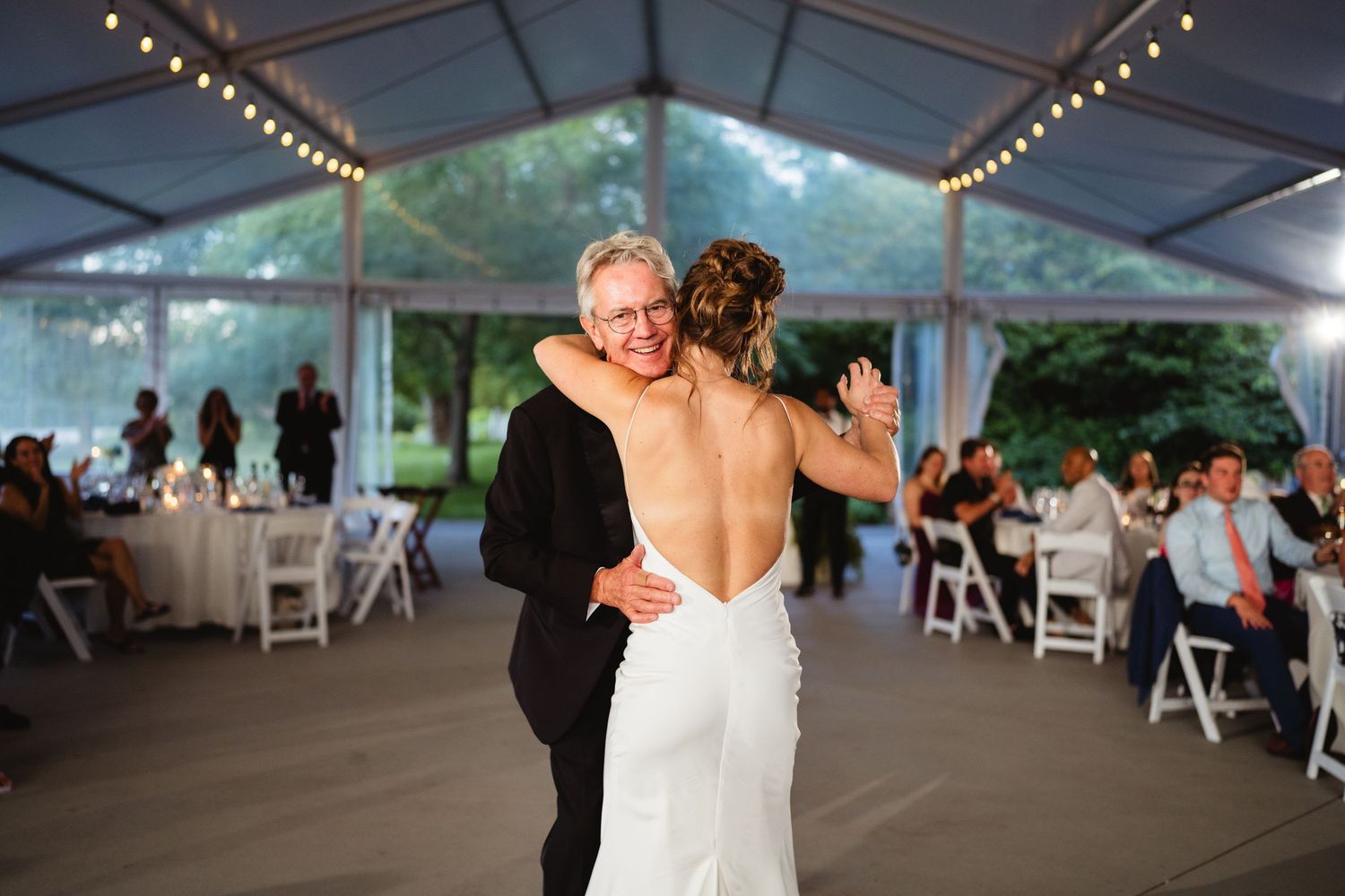 Bride and older man share an emotional first dance under a clear tent at an elegant outdoor wedding reception.