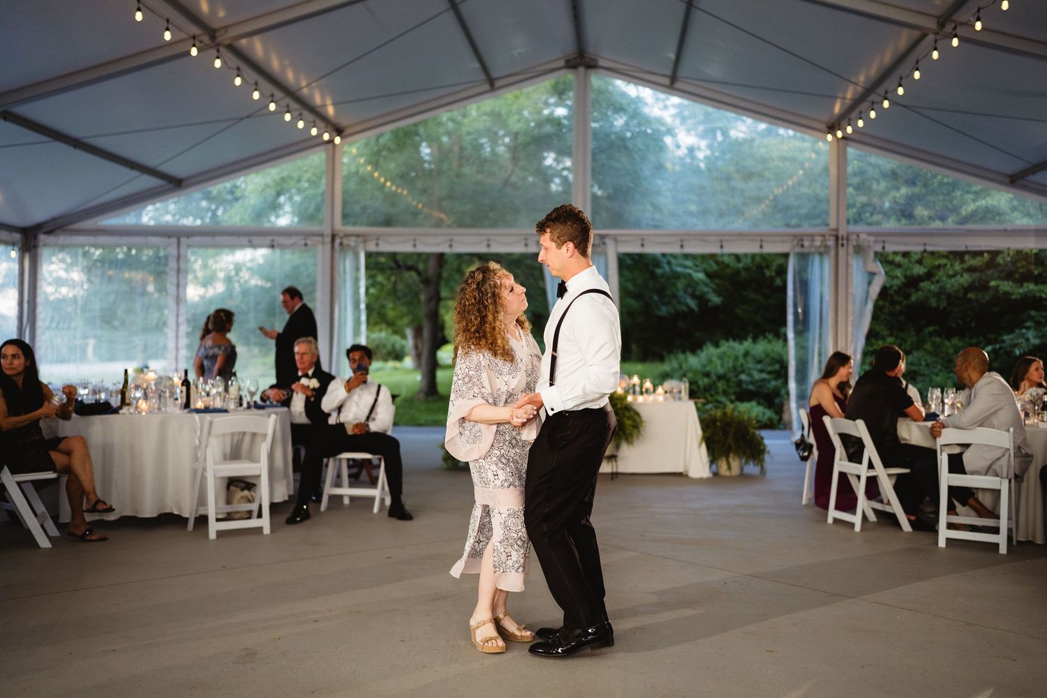 Couple shares a romantic dance under a clear wedding tent with guests seated at decorated tables nearby.
