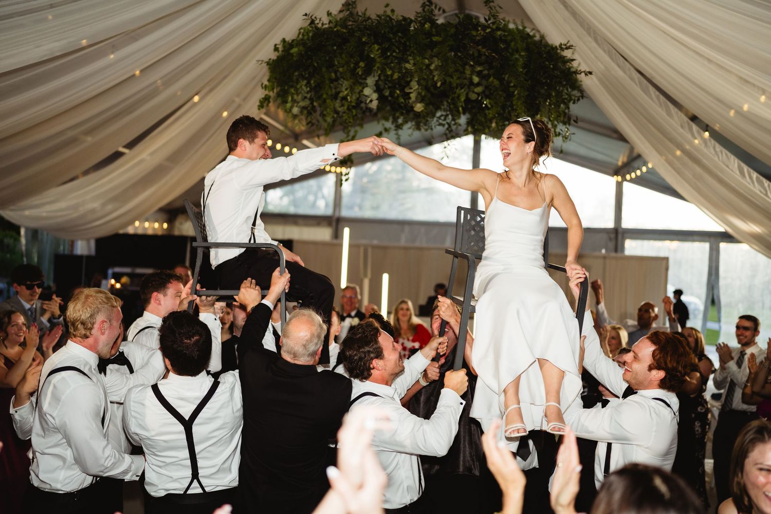Bride and groom lifted by groomsmen during joyful wedding reception hora dance under draped tent.