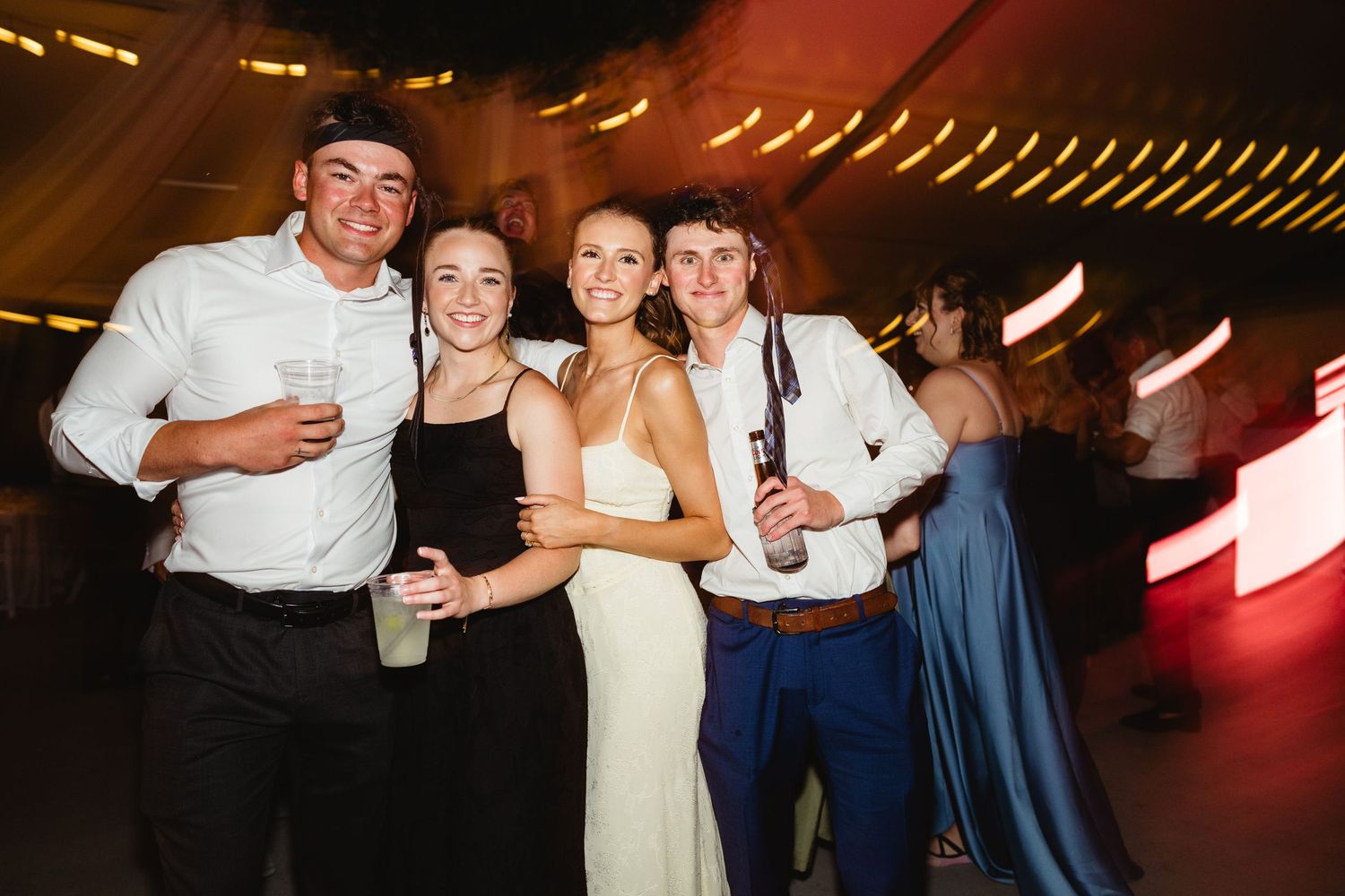 Four young adults smiling and posing together at a wedding reception party on a dance floor.