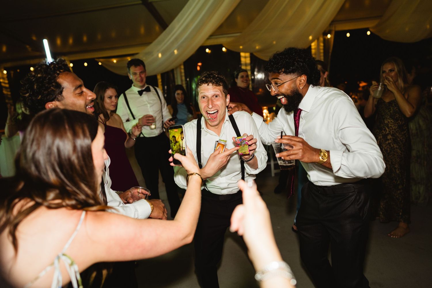 Guests dancing and celebrating at a lively wedding reception under a tent, holding drinks and smiling.