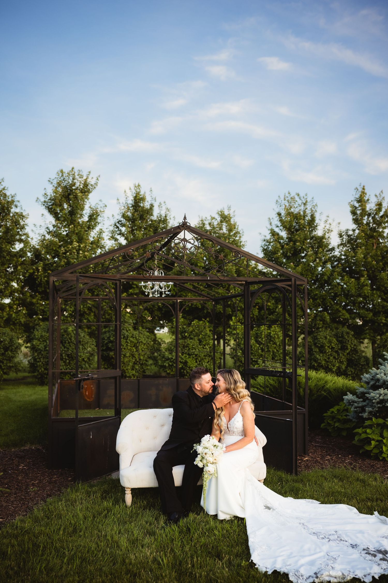 Bride and groom share a romantic kiss on a white sofa beside an elegant iron gazebo surrounded by lush green trees.