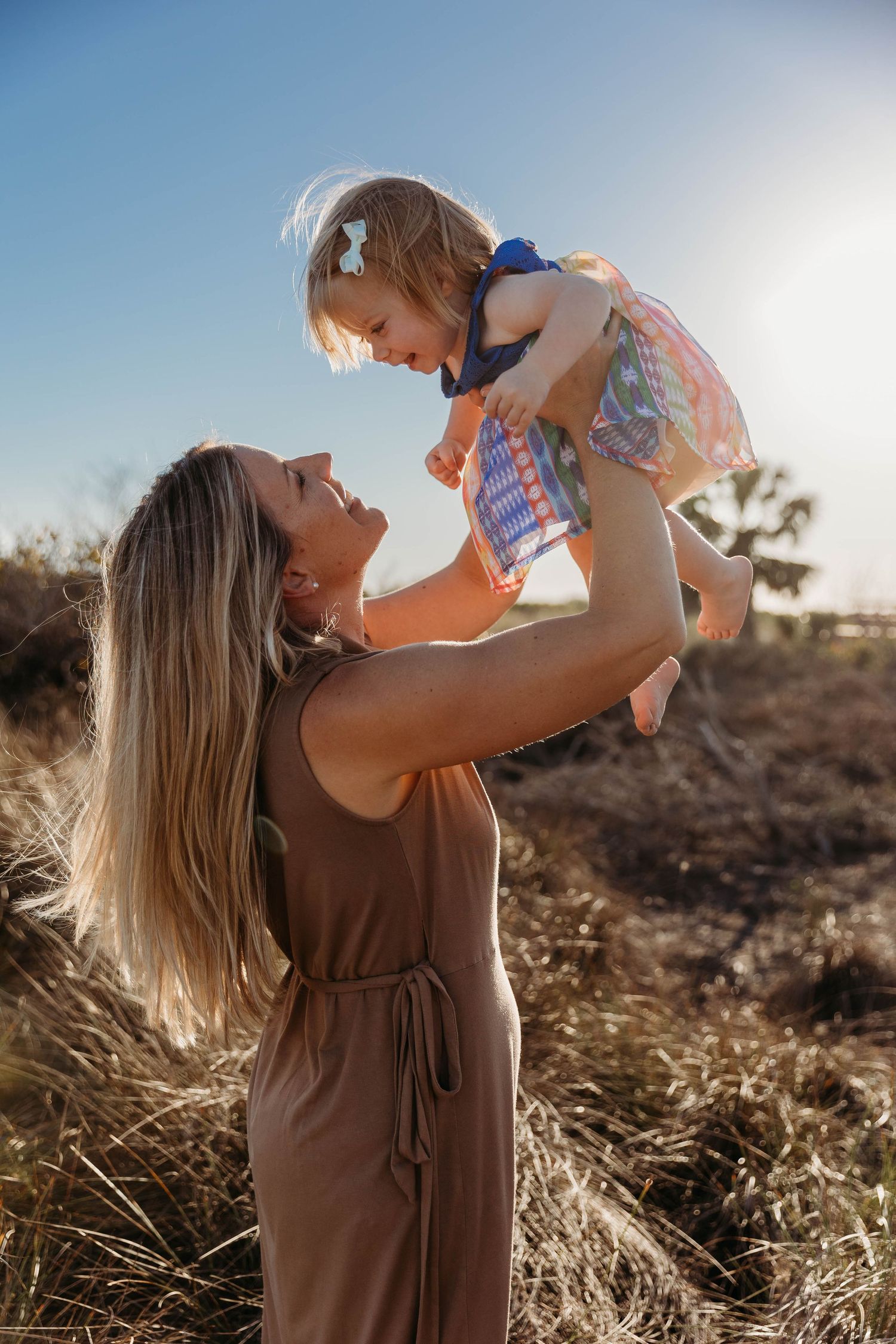 A mother joyfully lifts her toddler daughter in the air outdoors, both smiling in warm golden sunlight.