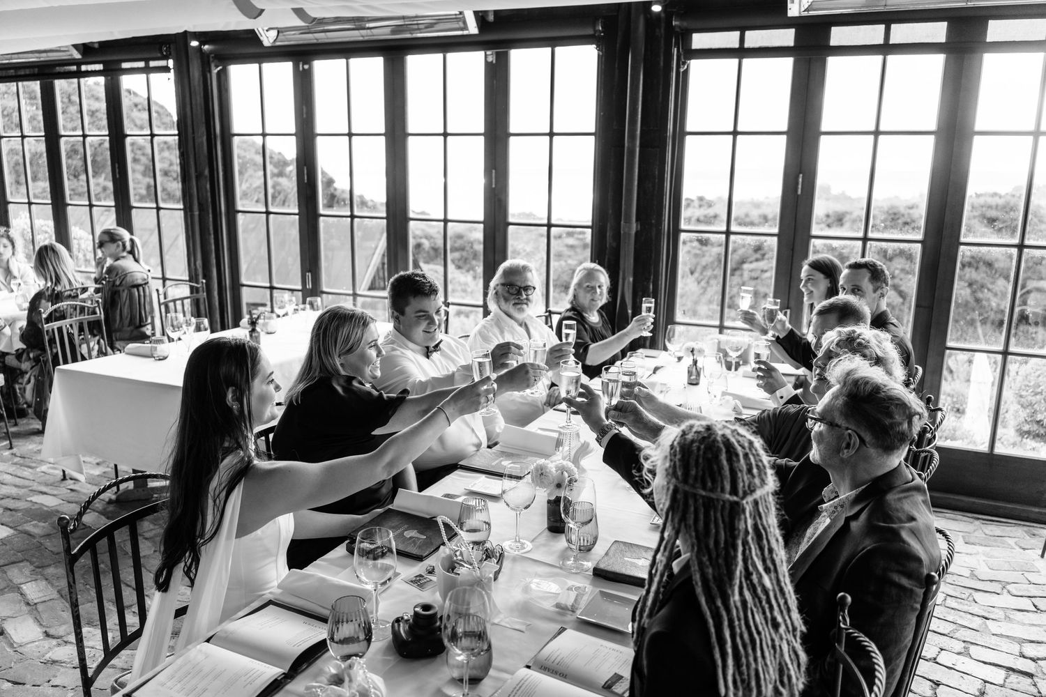 Black and white photo of wedding guests raising glasses in a toast at an elegant restaurant with large windows.