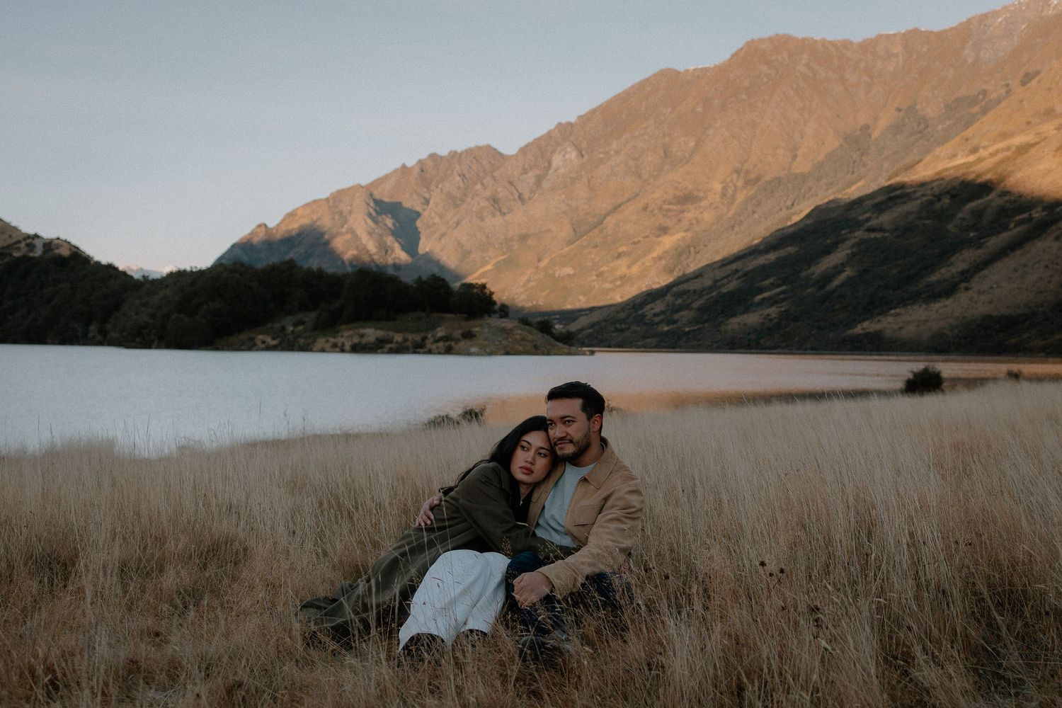 Couple embracing in golden grass field with mountain lake backdrop at sunset in New Zealand.
