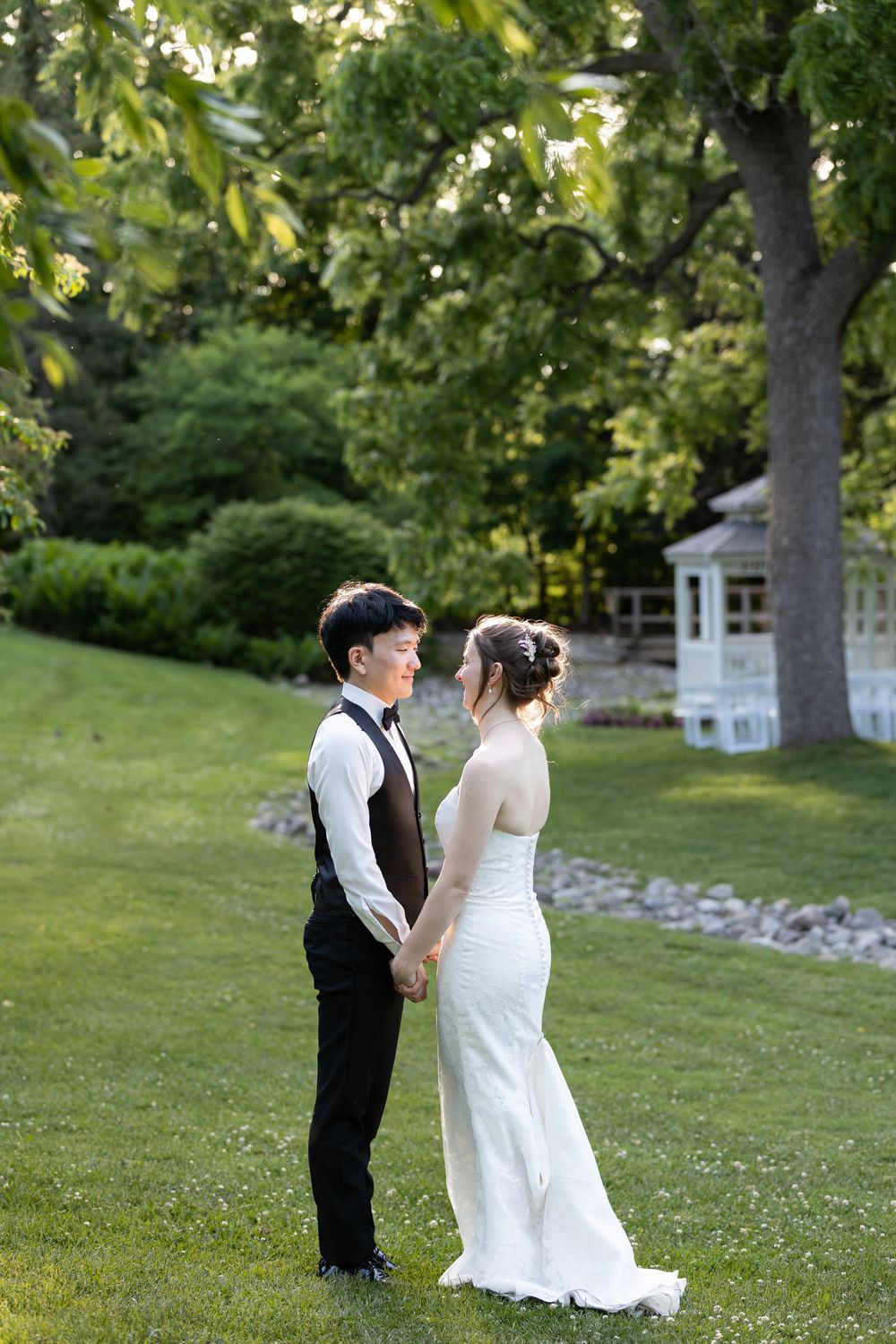 Bride and groom holds hands on a lush green lawn, with a white gazebo and trees during sunset at the civic garden complex in london, ontario