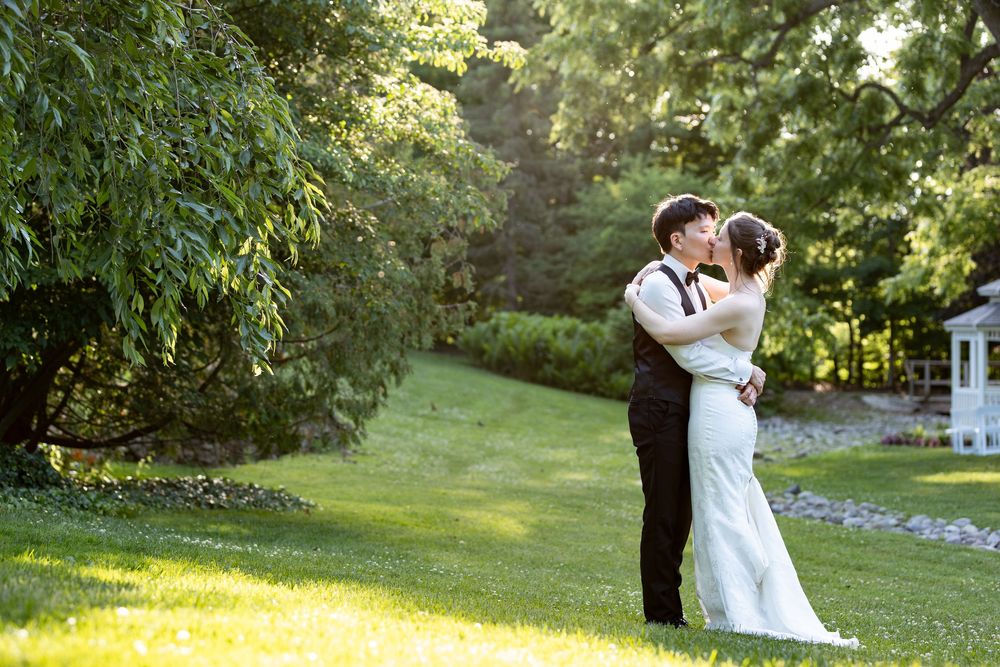 Bride and groom share a romantic kiss on a lush green lawn surrounded by trees during sunset at the civic garden complex in london, ontario.