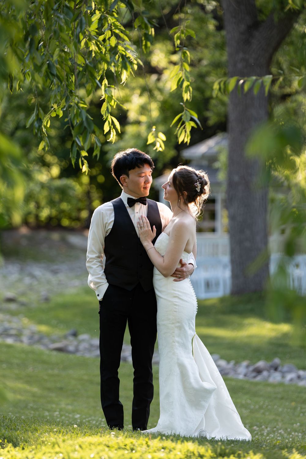 Bride & groom embrace under lush green trees in a sunlit garden during their wedding day at the civic garden complex in london, ontario.