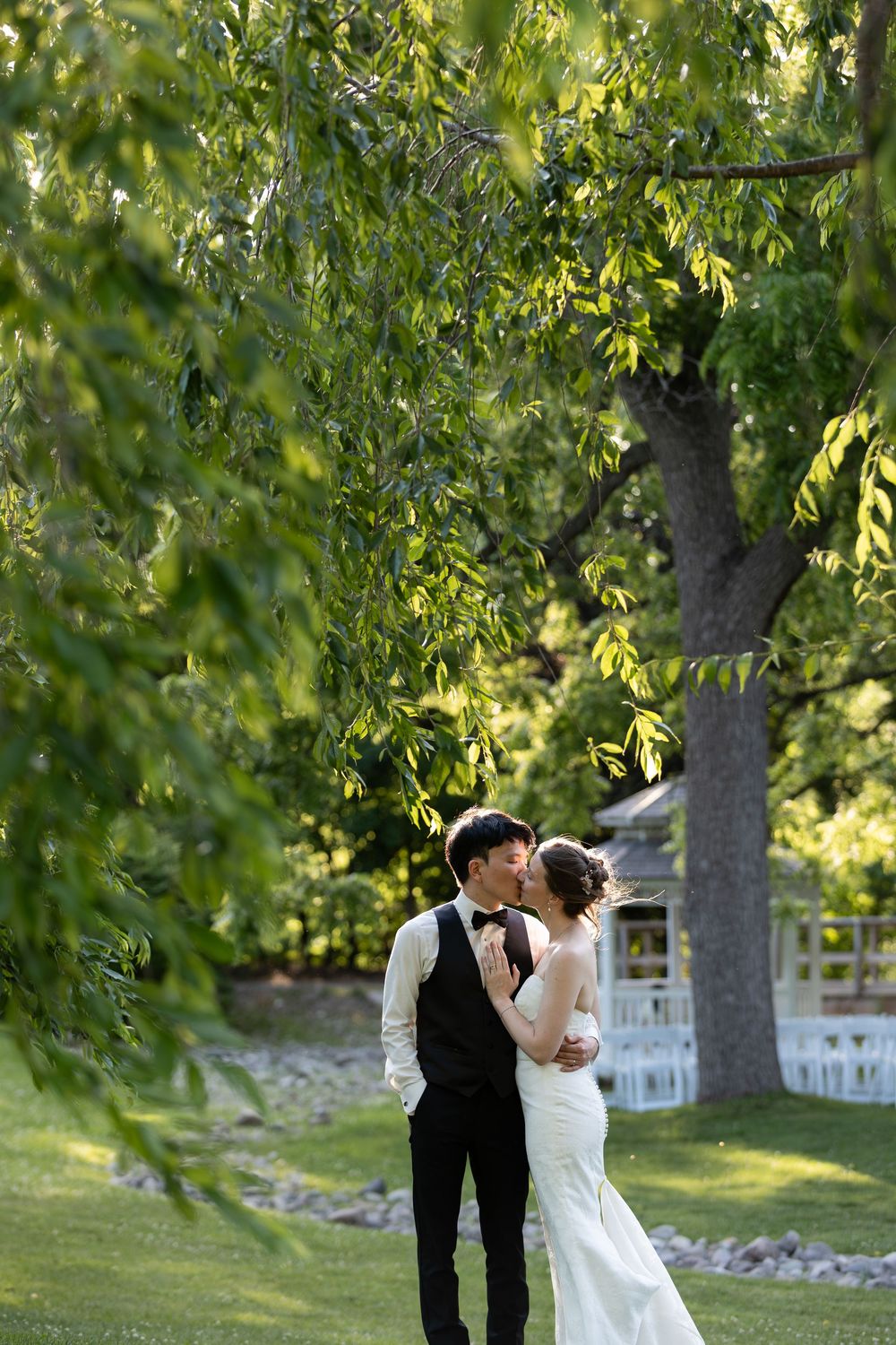 Bride and groom share a romantic kiss outdoors under lush green trees on their wedding day at the civic garden complex in london, ontario.
