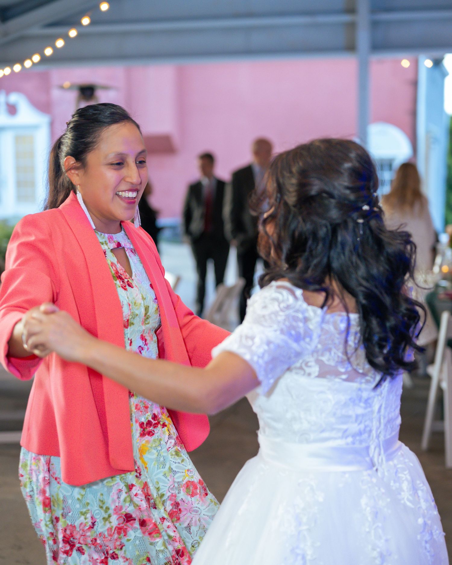 Two women dancing joyfully at a wedding celebration, one in a white gown and the other in a coral blazer.