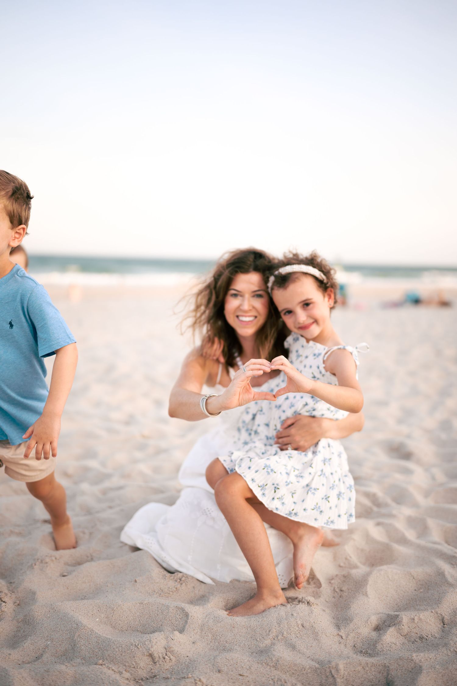 A smiling mother crouches on a sandy beach making a heart shape with her daughter in a floral dress.
