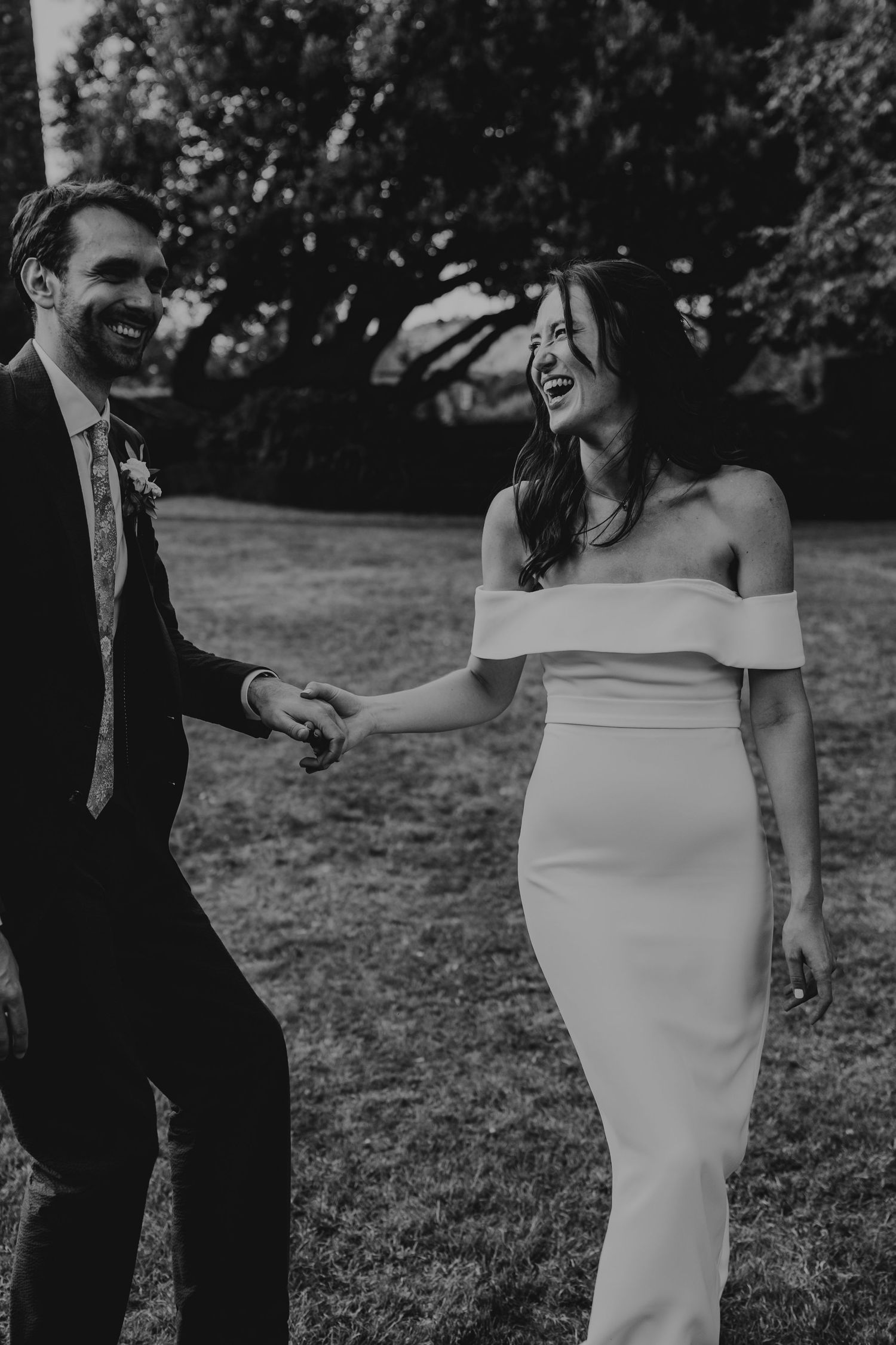 Black and white photo of a smiling bride in off-shoulder gown holding hands with groom in suit outdoors.
