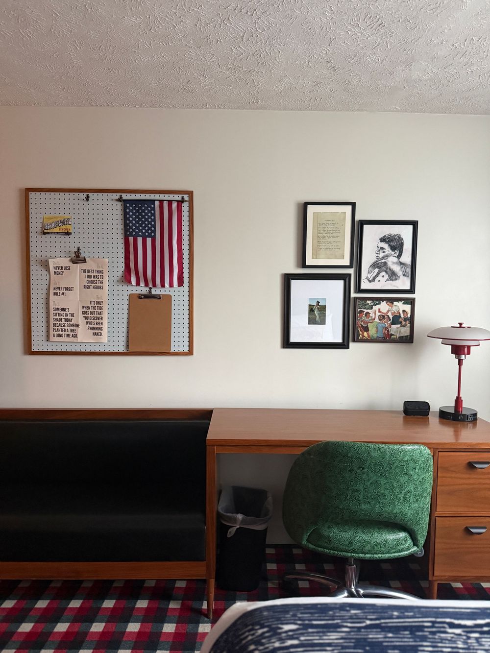 College dorm room with bulletin board, American flag, green chair, wooden desk, framed wall art, and plaid bedding.