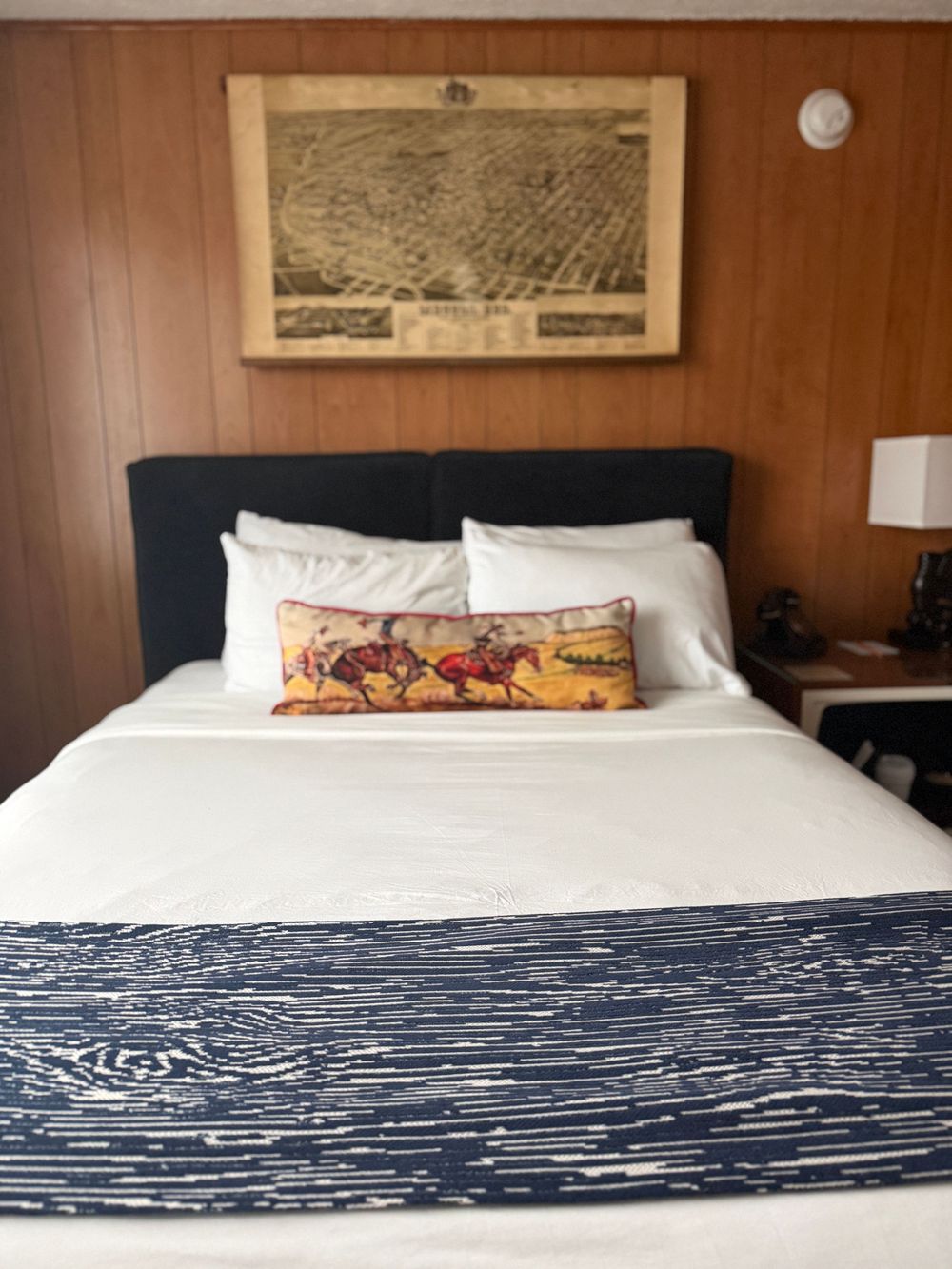 Cozy hotel bedroom with a king bed, navy runner, decorative horse pillow, and vintage city map artwork above.