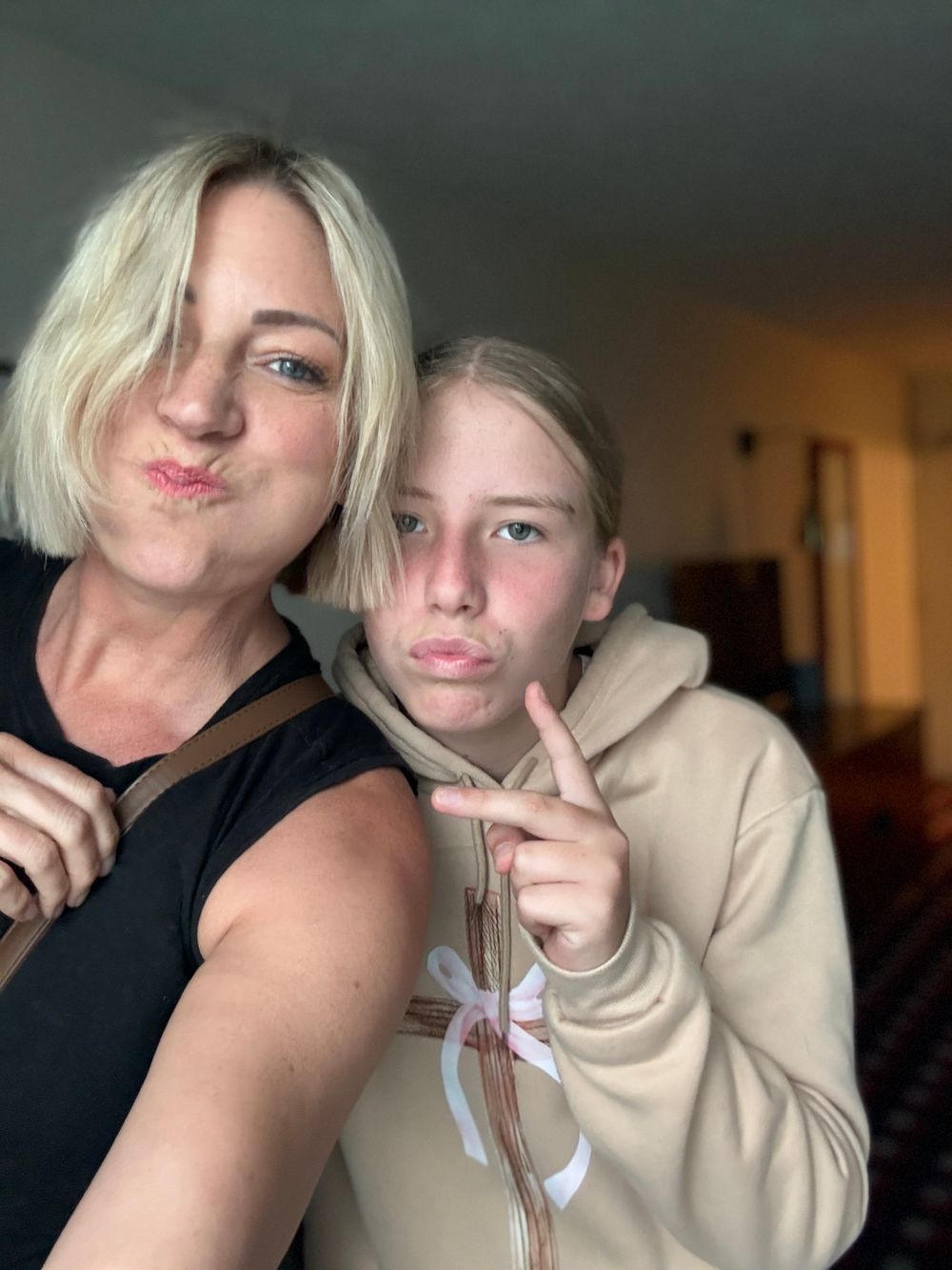 Two blonde females making funny faces, one flashing a peace sign, posing together for a selfie indoors.