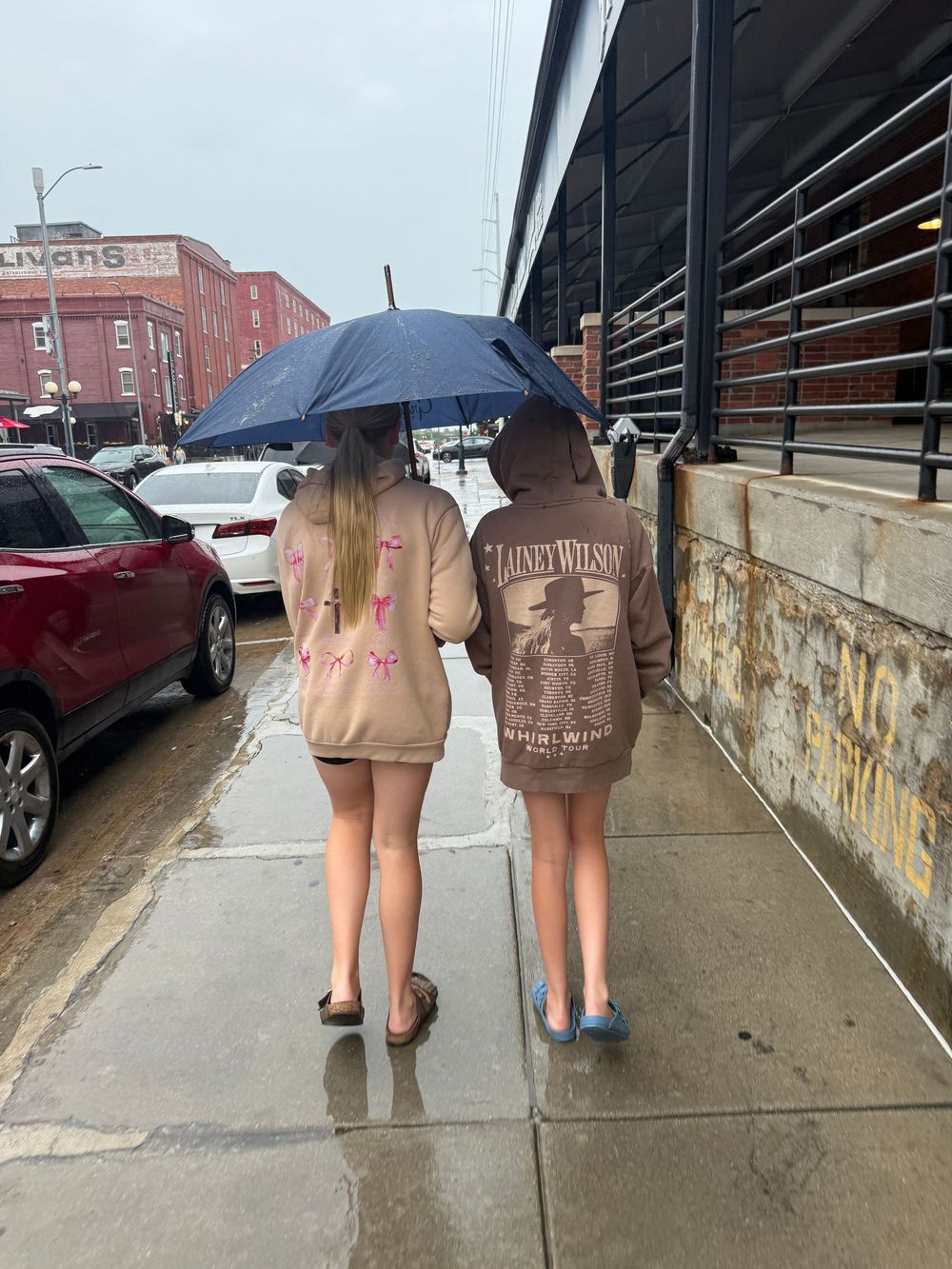Two girls sharing a blue umbrella on a rainy sidewalk wearing matching Lainey Wilson hoodies.