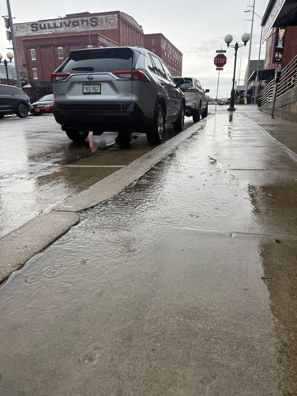 Flooded sidewalk next to parked SUV on wet urban street near Sullivan's sign.