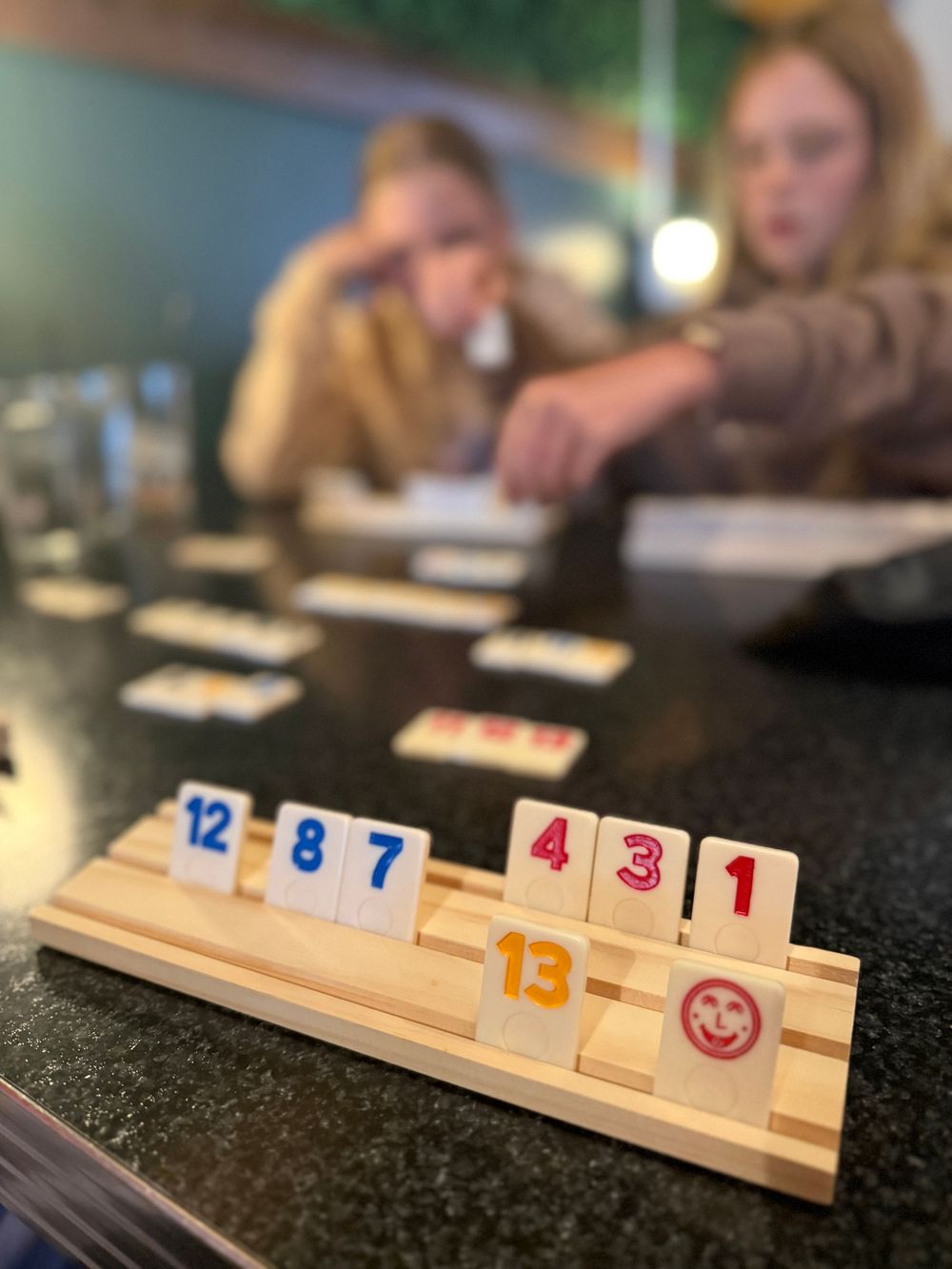 Two girls playing Rummikub board game with numbered tiles on a wooden rack at a table.