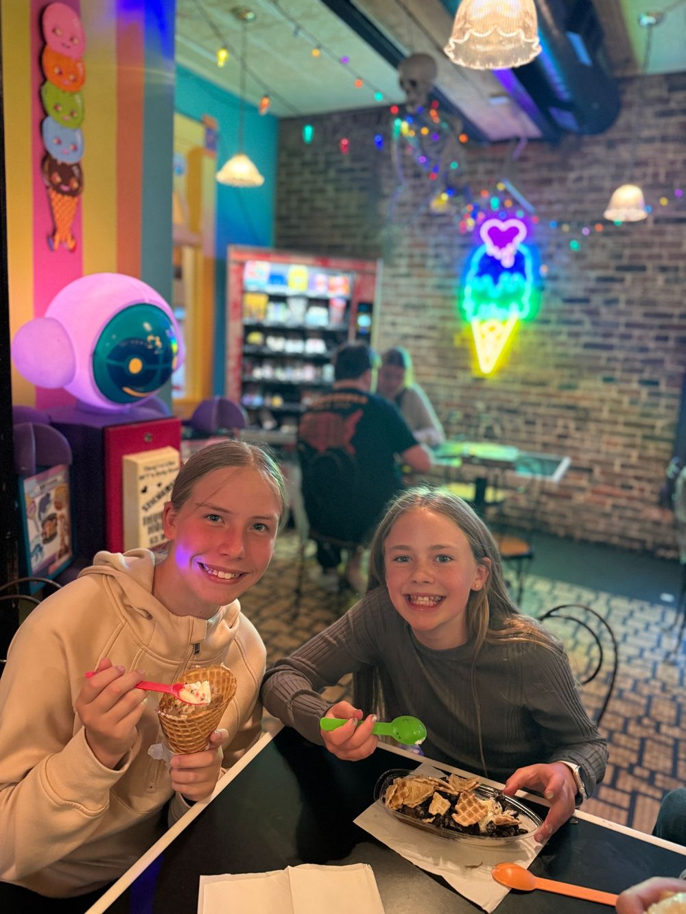 Two girls smile at camera in colorful ice cream shop with neon signs, one holding a waffle cone dessert.