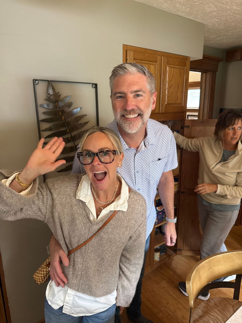 A cheerful woman with glasses waves while a smiling gray-haired man hugs her inside a cozy home.
