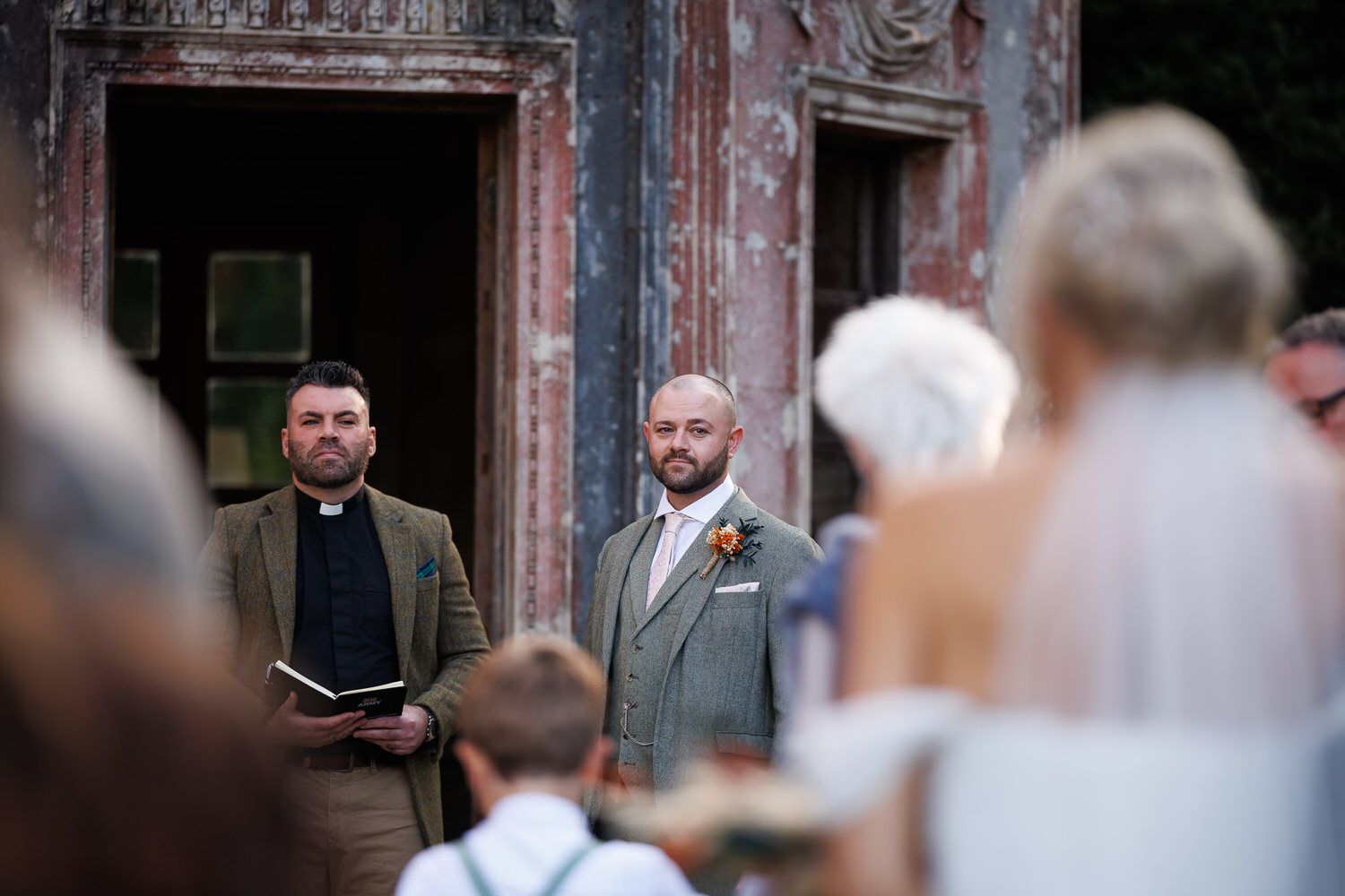 Groom in grey suit watches bride approach at outdoor wedding ceremony beside officiant holding book.