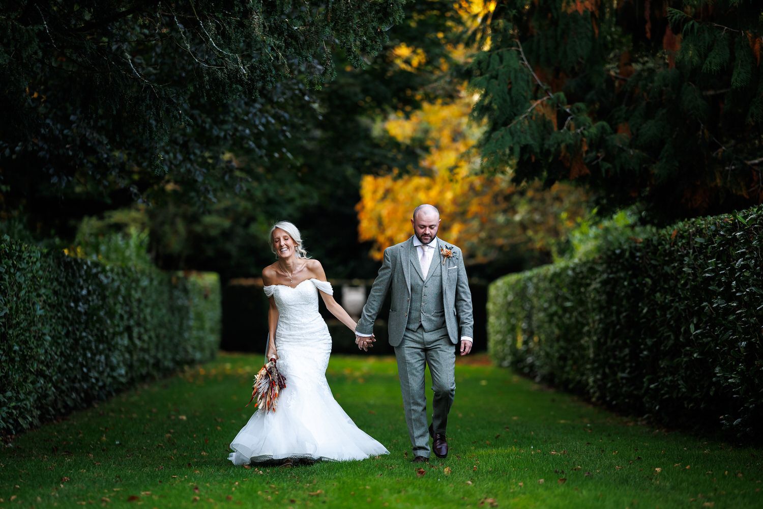 Bride and groom walking hand in hand through a lush green garden pathway on their autumn wedding day.