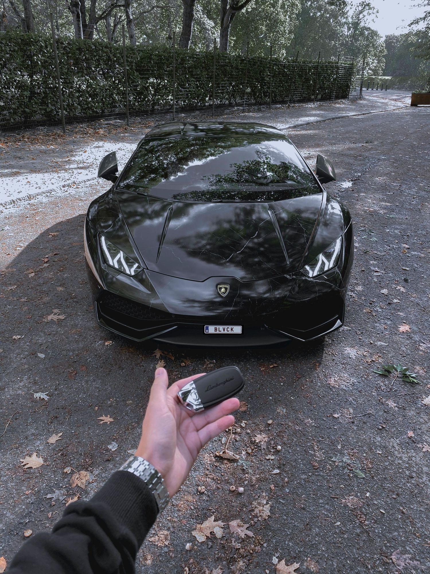 A person holds a black Lamborghini Huracan key fob in front of a sleek matte black Lamborghini sports car parked outdoors.