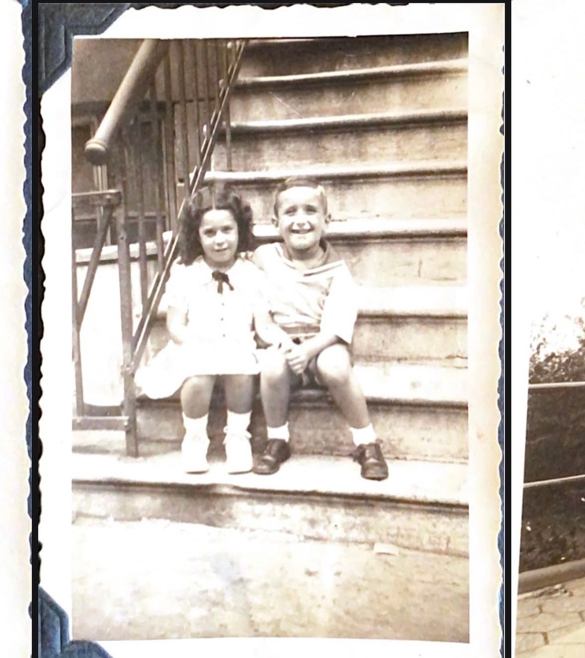 Vintage black and white photo of two young children sitting together on outdoor stone steps, smiling at the camera.