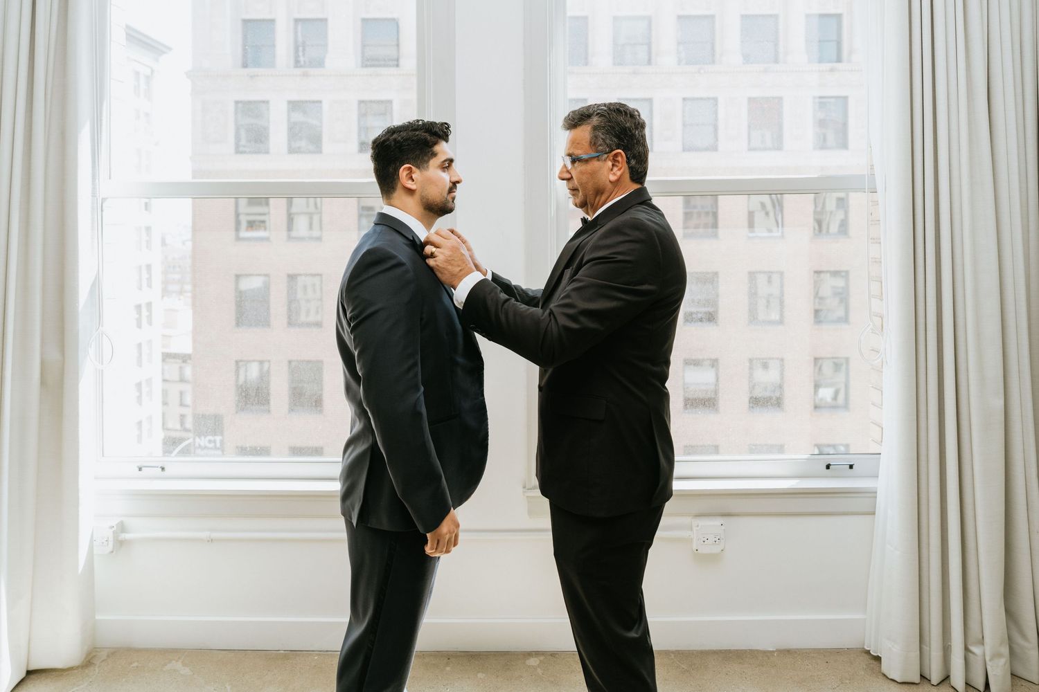 Groom standing with his father in front of large windows during final preparations.
