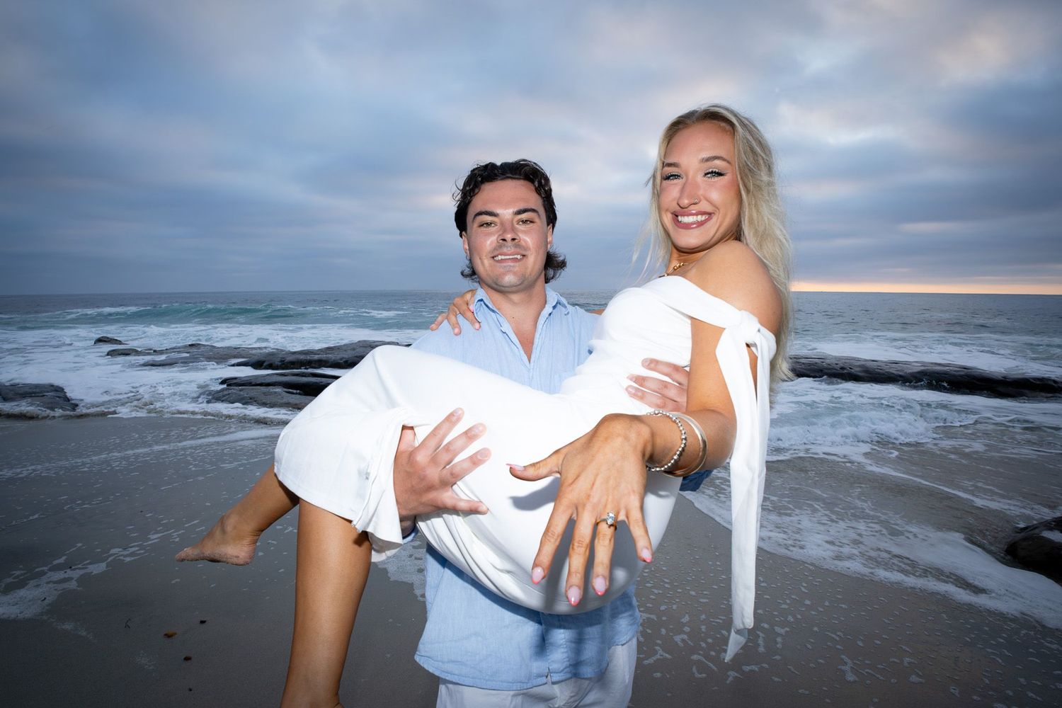 San Diego engagement photography of a man lifting a woman on a beach as she shows off her diamond surprise proposal ring.