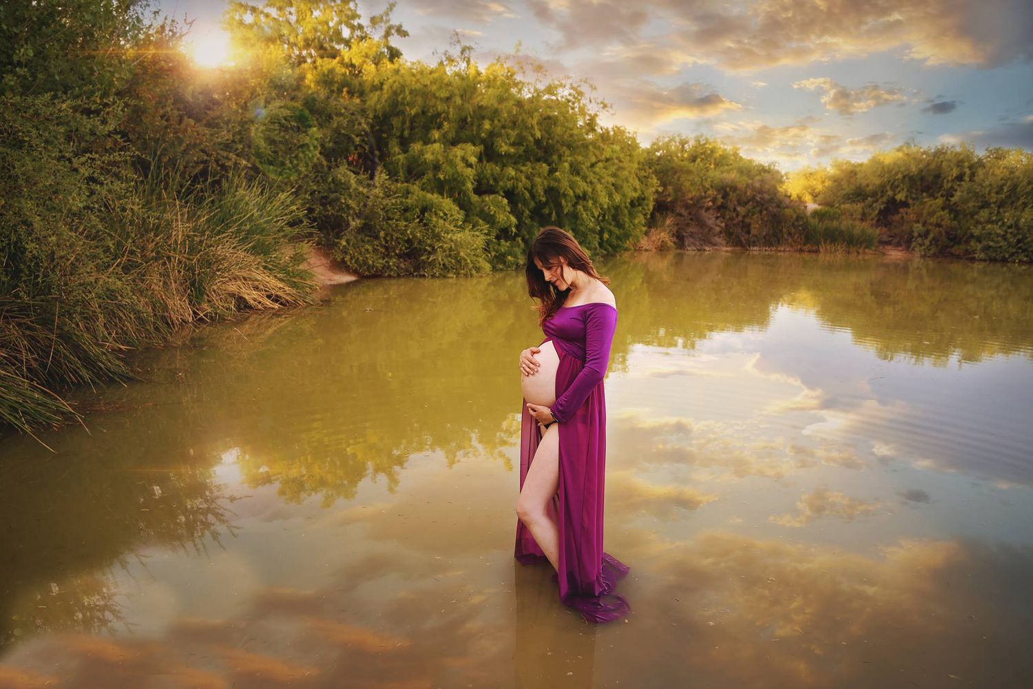 Pregnant woman in a magenta maternity gown stands in shallow water at sunset, with golden clouds reflected on the calm surface.