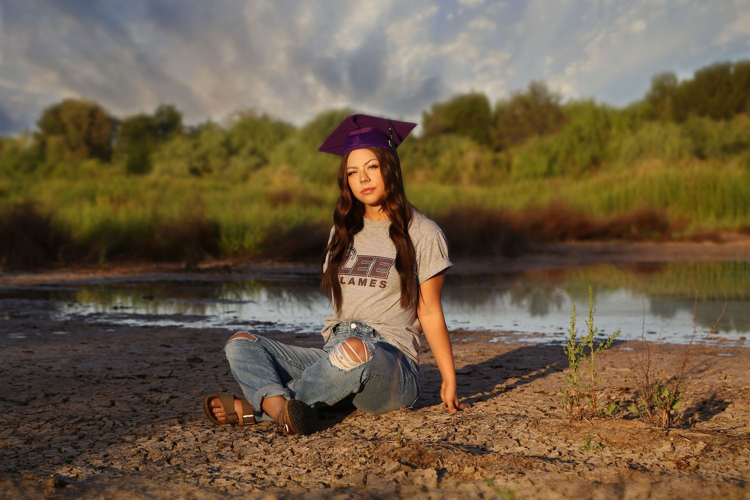 Young woman in a purple graduation cap and graphic tee sits on a muddy shoreline with green trees behind her at sunset.