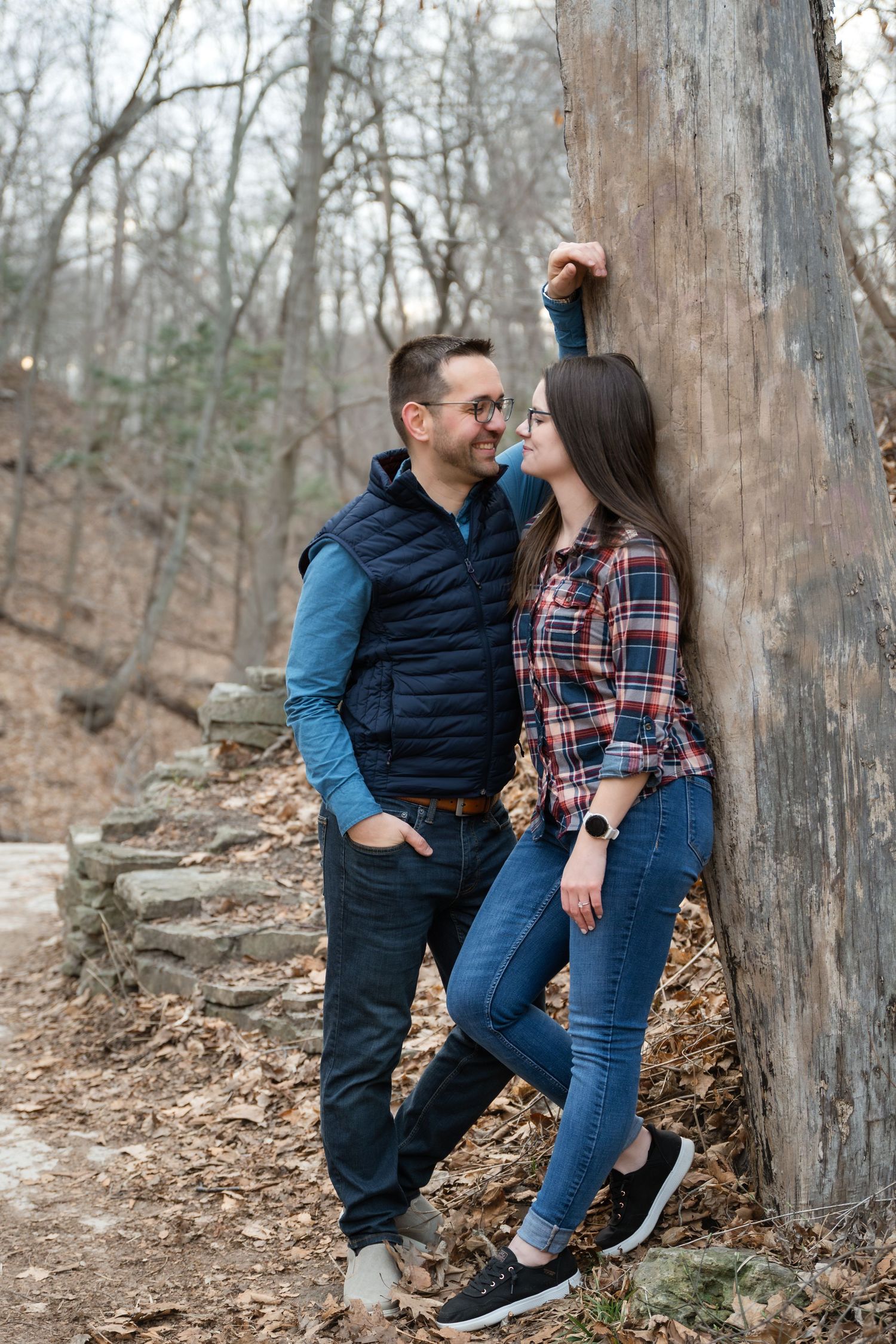 Couple sitting on stone steps in autumn forest, woman in plaid shirt and glasses leaning on man in blue vest.