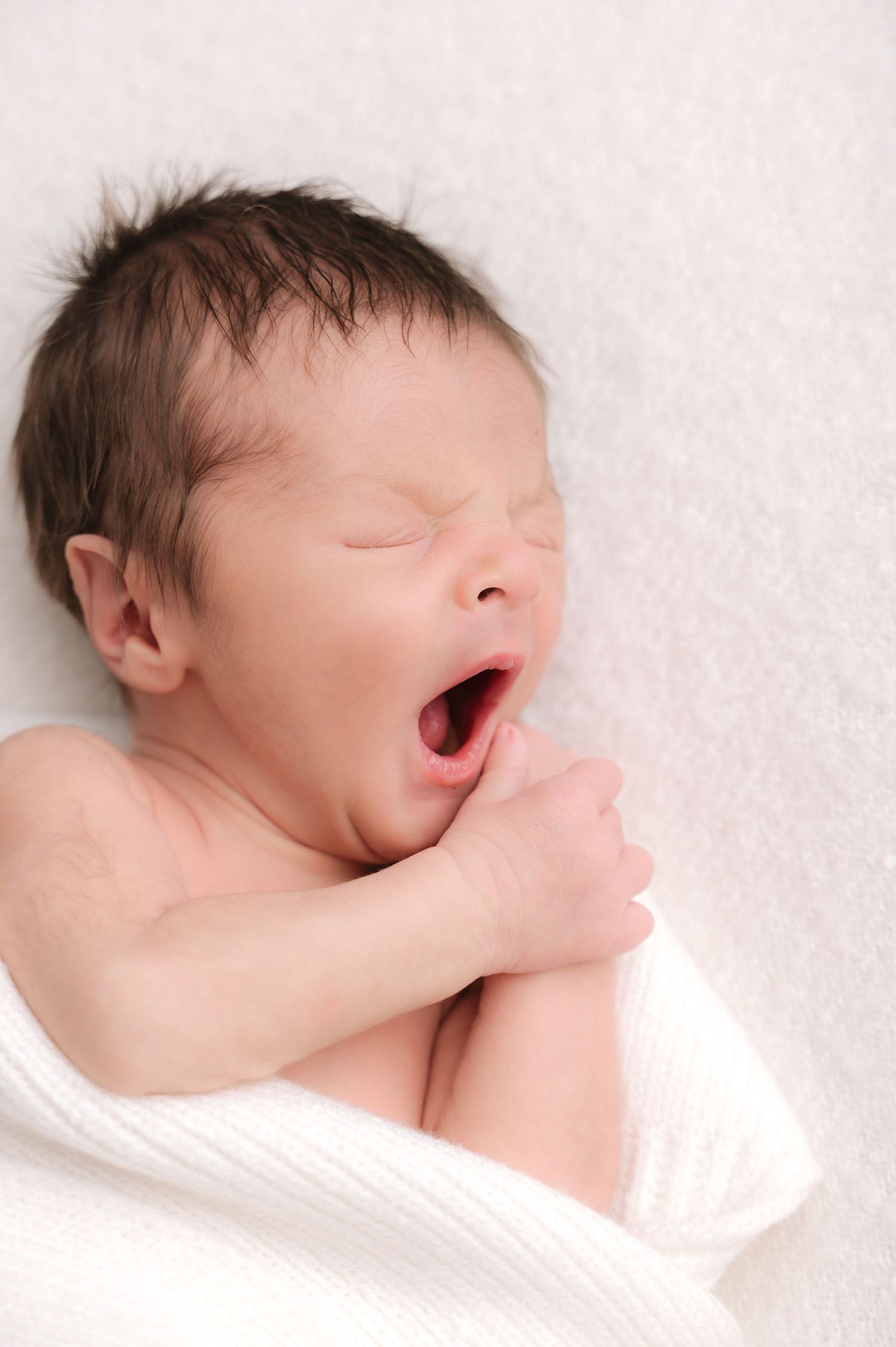 Newborn baby yawning while swaddled in a white blanket, with eyes closed and tiny fists raised to chin.