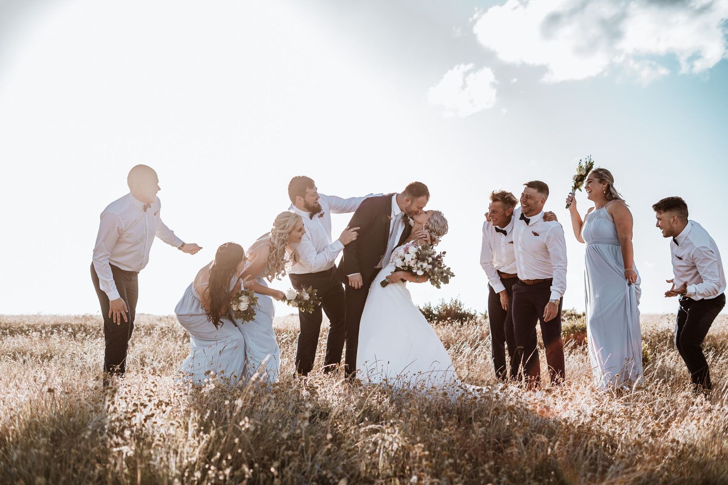 Wedding party celebrating in golden field with bride and groom kissing under bright sunny sky.