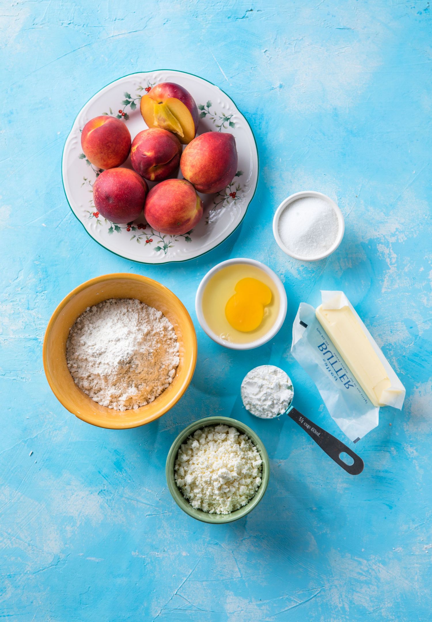 Fresh peaches, flour, butter, egg, sugar, and cornstarch ingredients laid out on a blue surface for a peach dessert recipe.