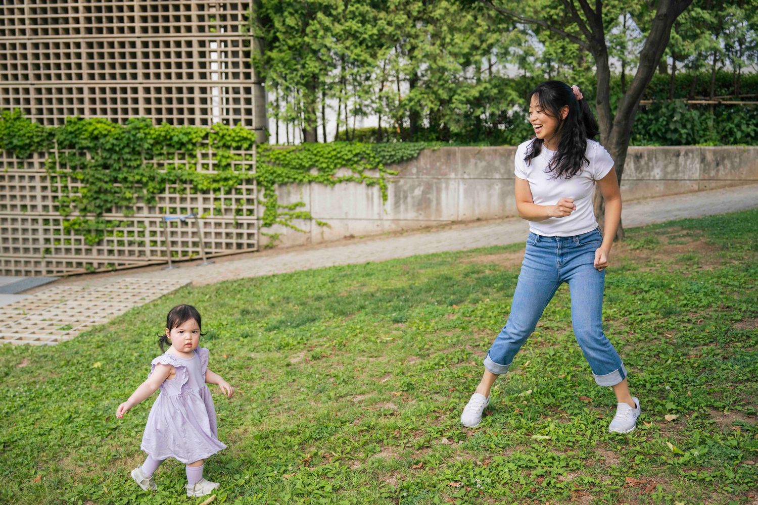 A mother and toddler daughter playing together on green grass in a sunny park setting.