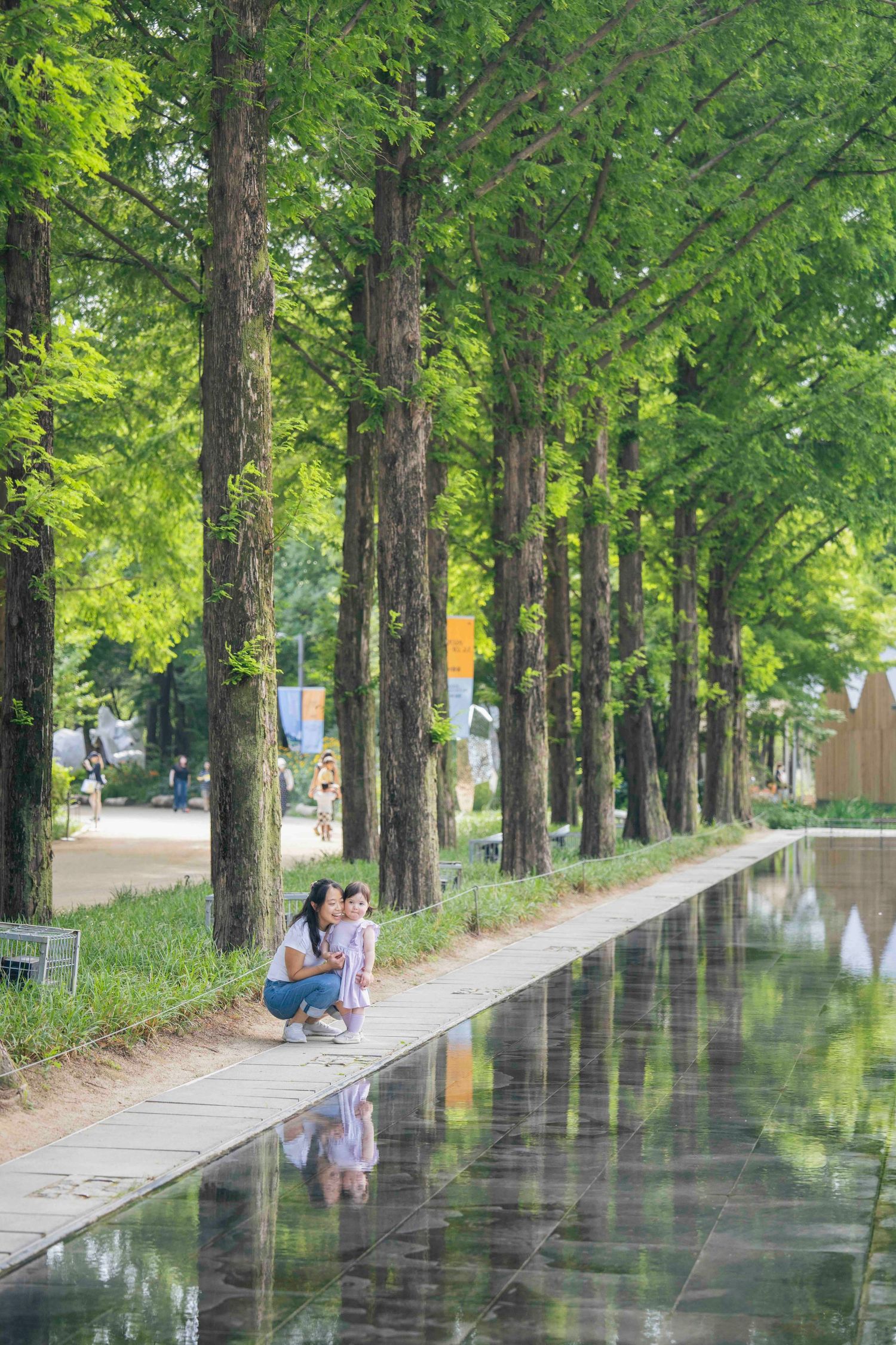 Mother and child crouching by a reflective pool along a tree-lined path in a lush green park.