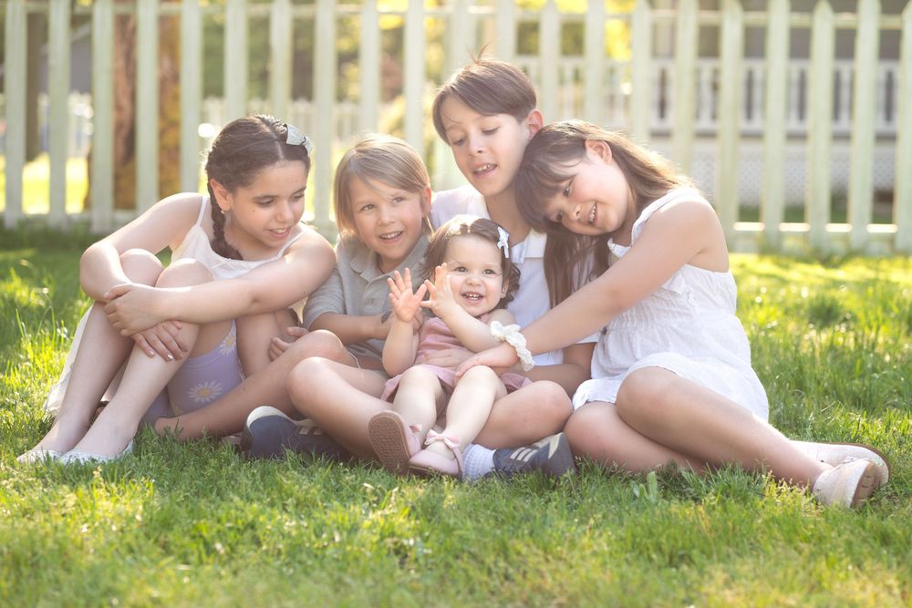 Five children sitting together on green grass in a sunny backyard, smiling and hugging near a white picket fence.